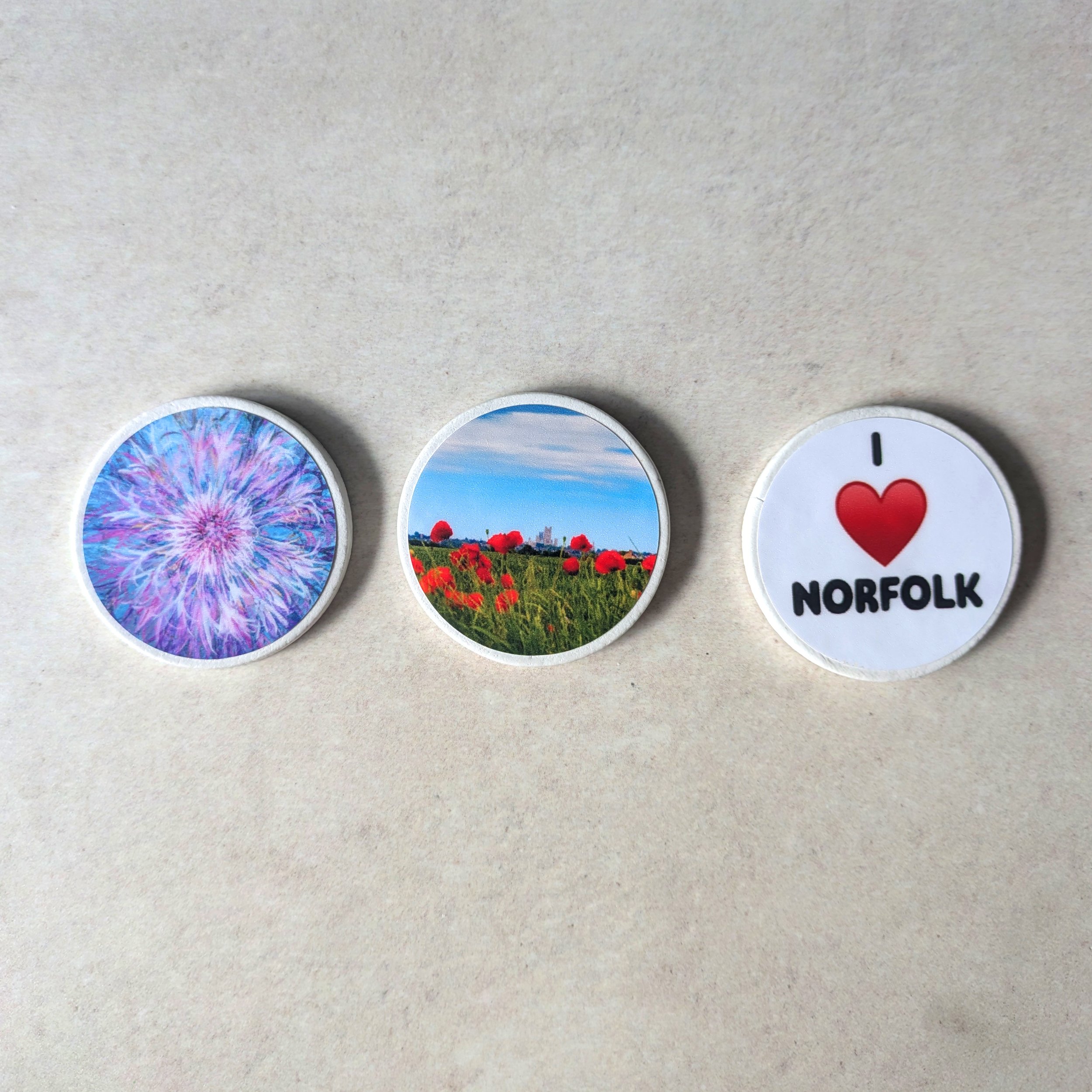 Three custom-made round fridge magnets displayed in a horizontal row on a neutral background. From left to right: a purple mandala design, a landscape photo of red flowers, and the 'I LOVE NORFOLK' heart design. Handmade by OH Arts in the UK.