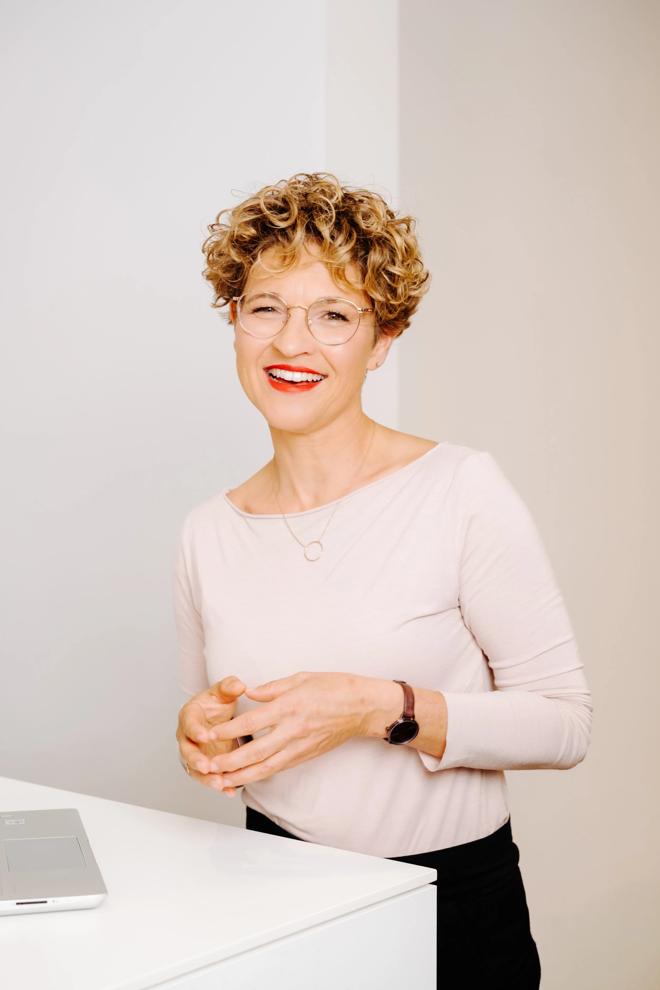 Lächelnde Frau mit Kurzhaarfrisur, Brille, rotem Lippenstift und weißem Oberteil, steht vor weißer Wand neben Laptop.