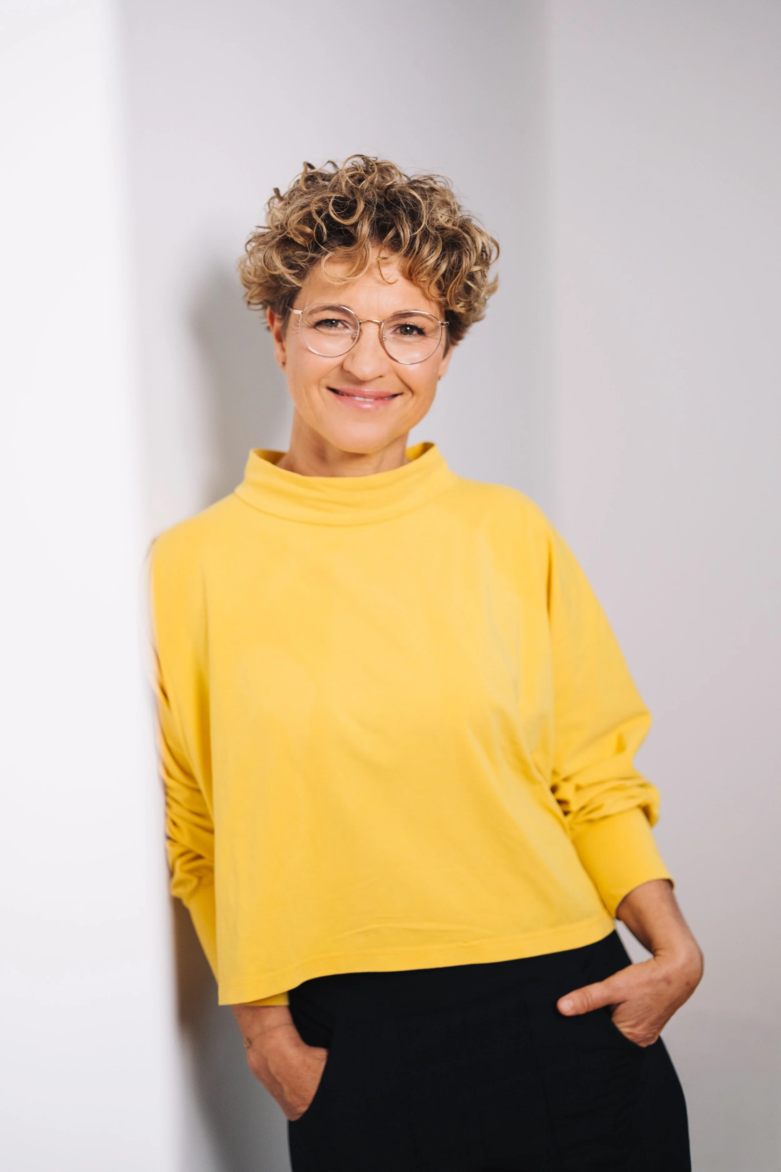 Eine lächelnde Frau mit kurzen, lockigen Haaren trägt eine gelbe Sweatshirt und schwarze Hosen, steht vor weißer Wand.