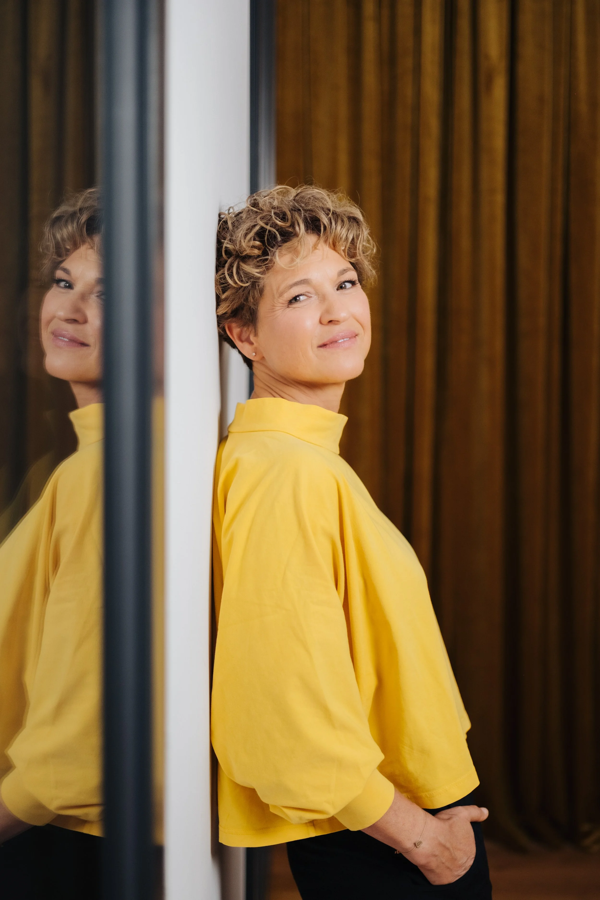 Eine Frau mit kurzen, lockigen Haaren trägt eine gelbe Bluse und lehnt an einer Wand, während sie in eine Kamera lächelt. Ihre Spiegelung ist im Glas auf der linken Seite sichtbar.