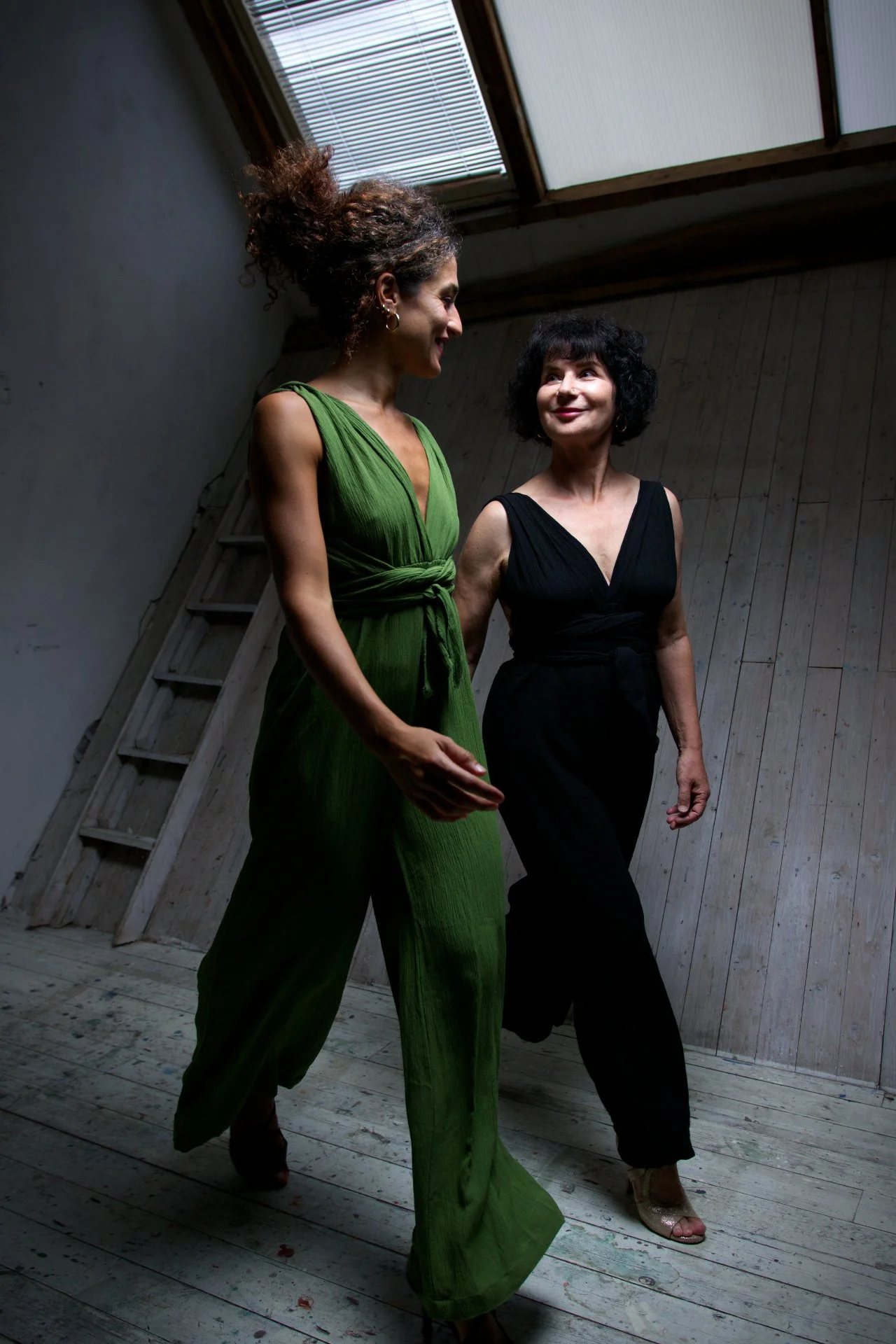 Two women walking together in a wooden room with a slanted ceiling and skylight, one in a green dress and the other in a black dress, smiling at each other.