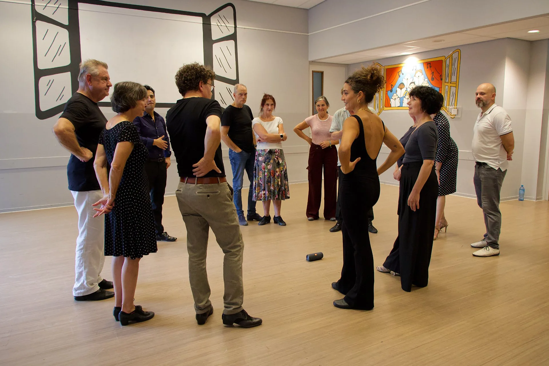 A group of people gather in a circle in a room with wood flooring and colorful murals on the walls, engaging in a discussion or activity.