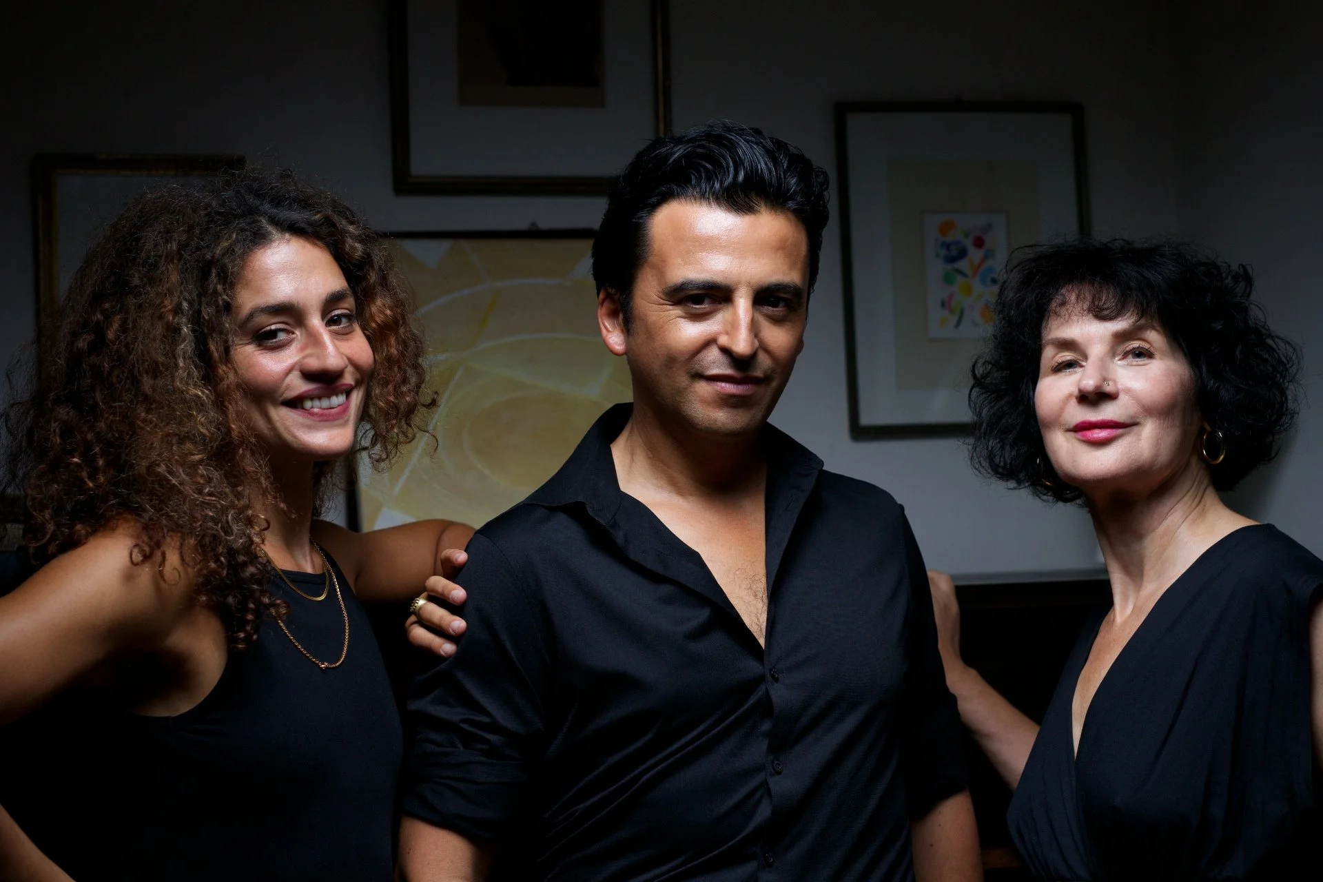 Three people standing close together, smiling, with framed artwork on the wall behind them.