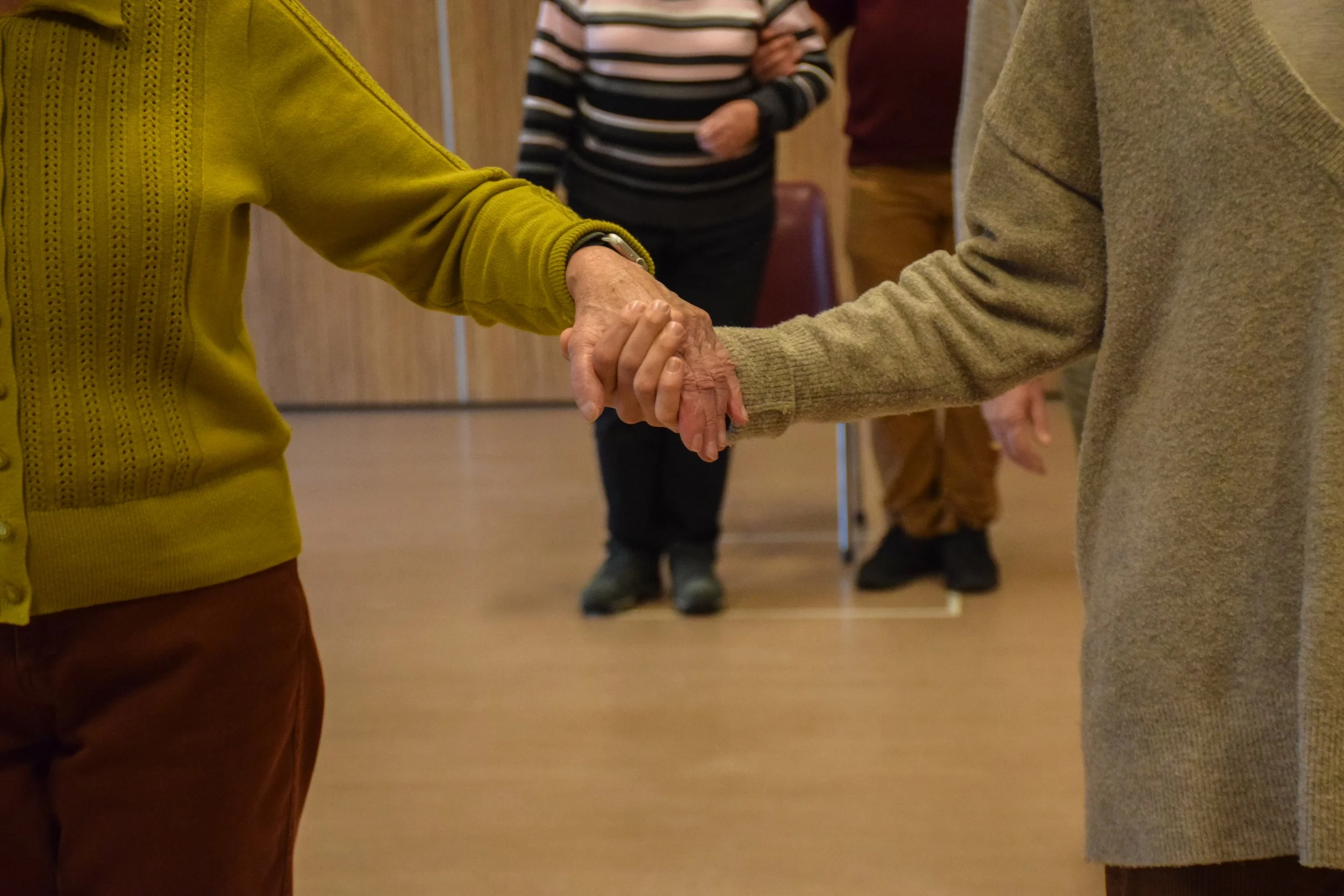 Two elderly people holding hands during a dance or social activity in a community space, with other individuals in the background.