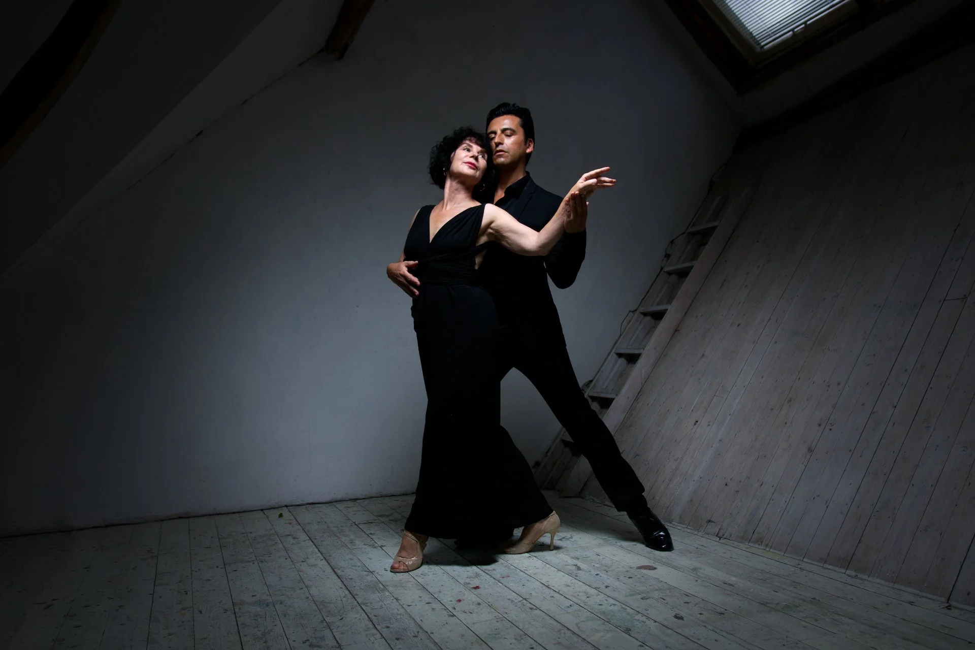 A man and woman dance closely together in a dimly lit room with slanted walls and wooden floors, the woman in a black dress and the man in black attire.