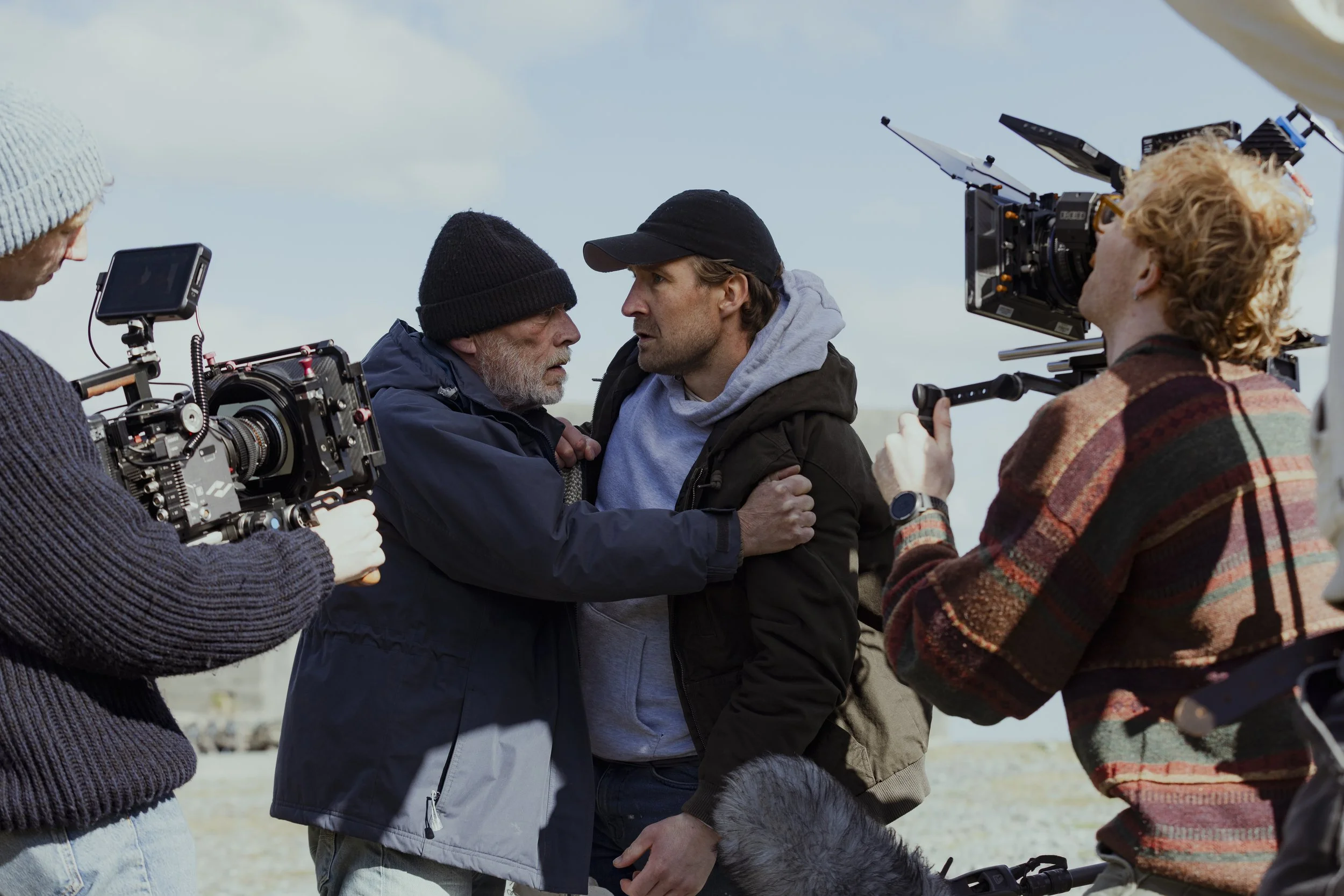 Scene from a film set showing two men in an intense confrontation, surrounded by a director and camera crew filming outdoors.