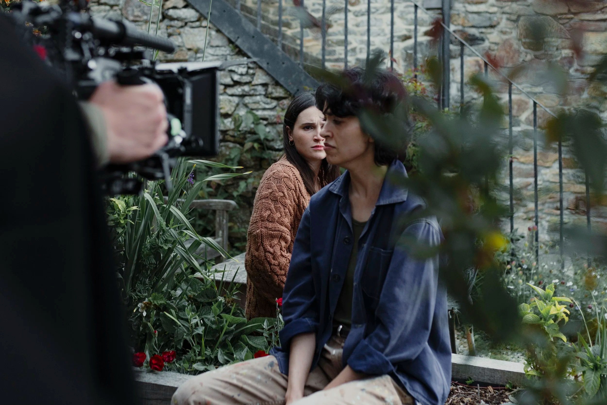 Two women outdoors in a garden with greenery and flowers, one with dark hair and a brown sweater looking concerned, the other with black hair and a blue jacket sitting on a ledge, with a person holding a camera on the left capturing the scene.