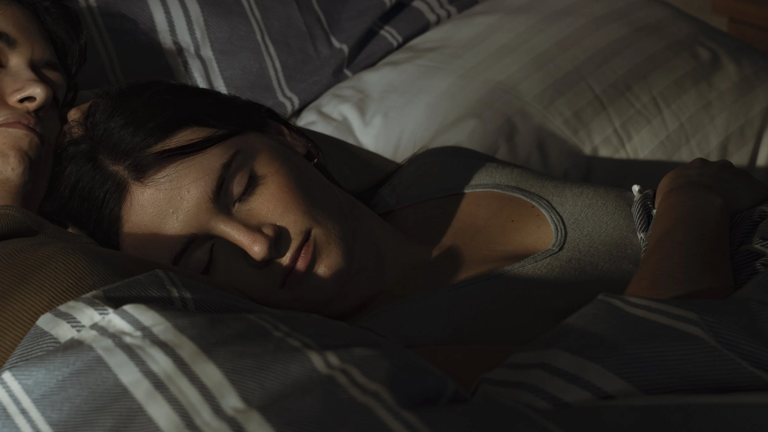 A woman sleeping peacefully in bed with a man close by, both surrounded by striped and plain pillows and bedding.