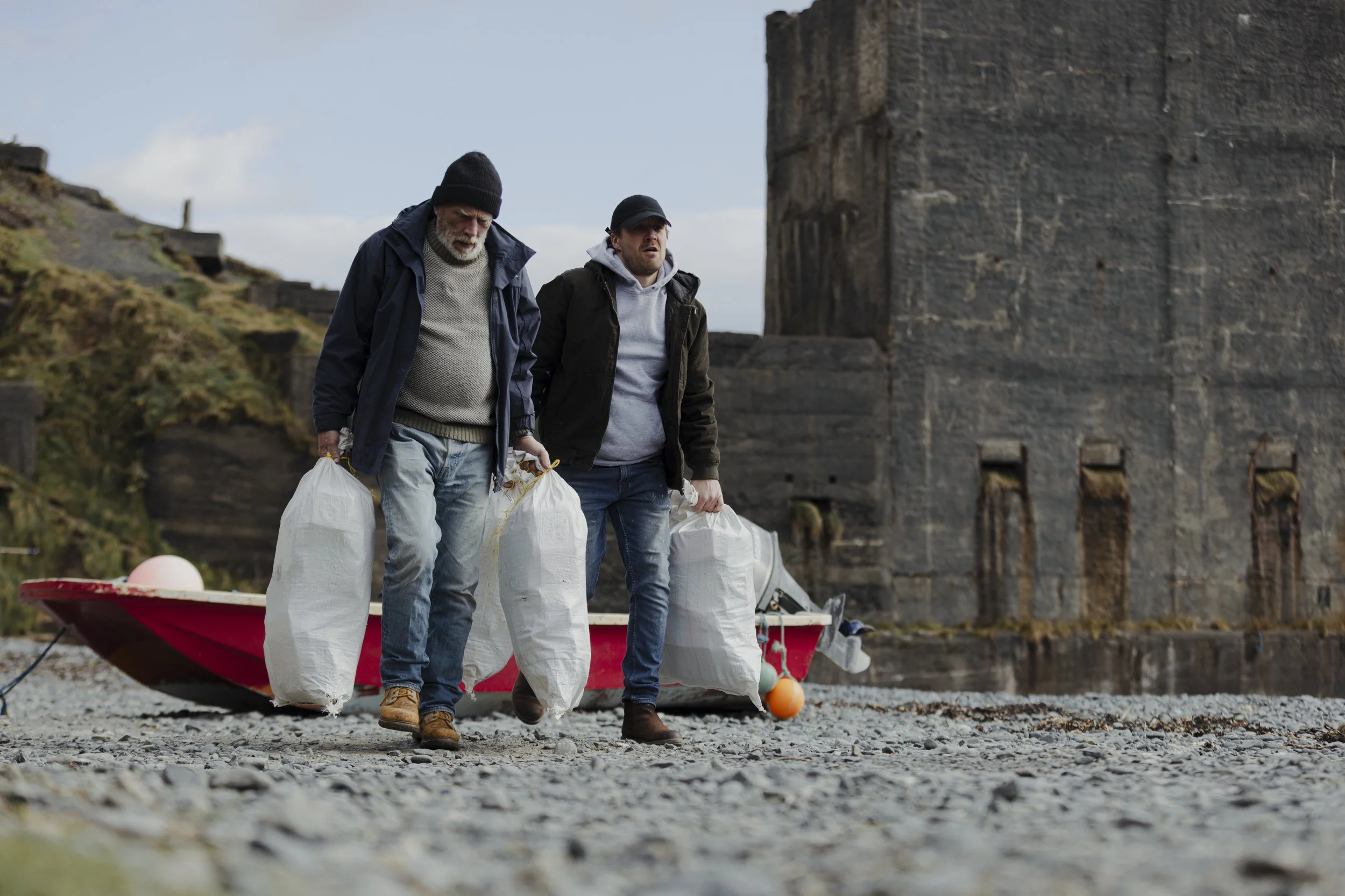 Two men walk along a rocky shore carrying white bags, with a red boat and a historic stone building in the background.