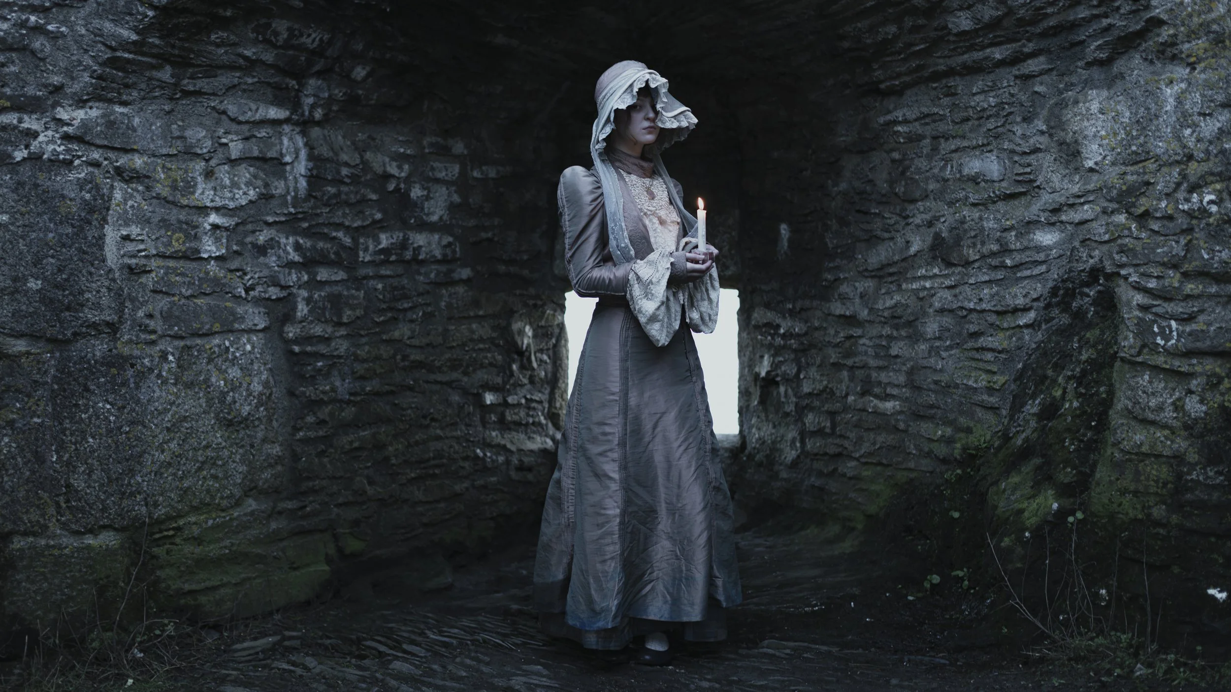 A woman dressed in historical or fantasy clothing, holding a lit candle, stands inside a stone archway or tunnel with moss and plants growing on the rocks.