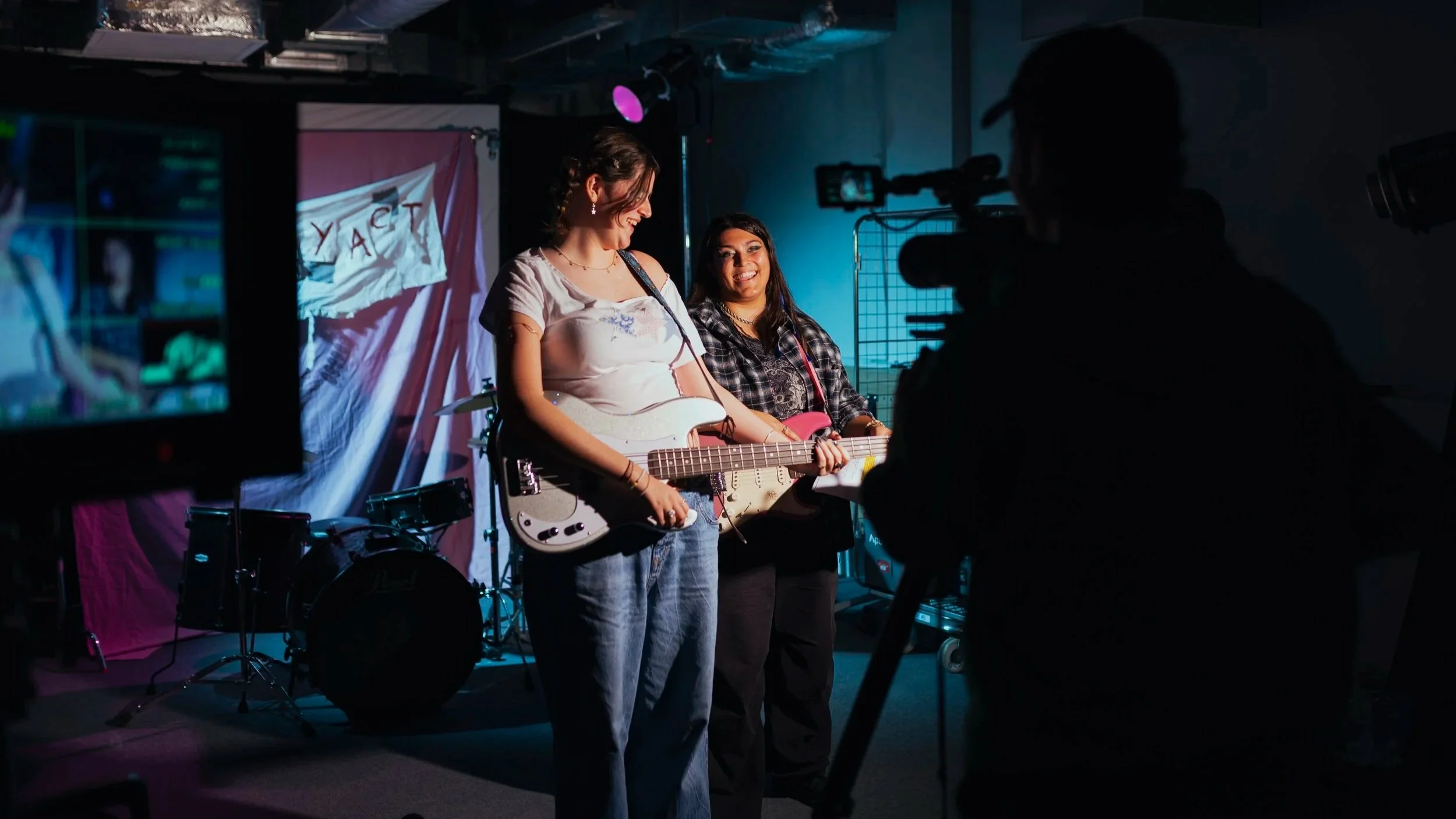 Two women with guitars being filmed in a music studio with a drum set in the background and a red, white, and pink curtain, on a dark room with studio lighting.
