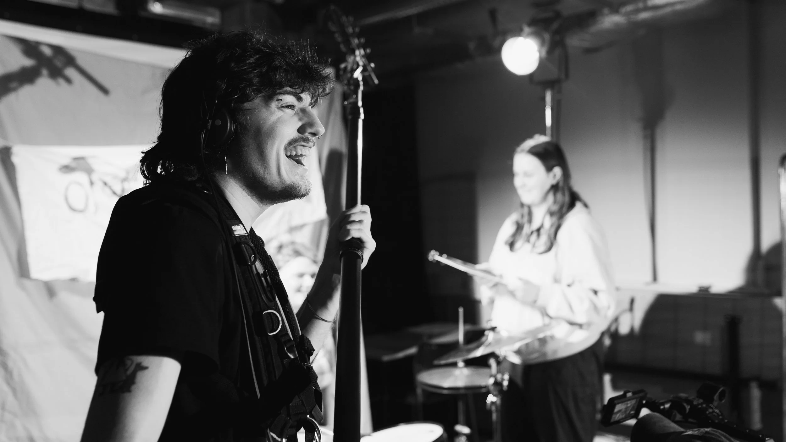 Black and white photo of a man with headphones smiling and holding a microphone stand, a woman in the background reading a script and playing drums in a recording studio.