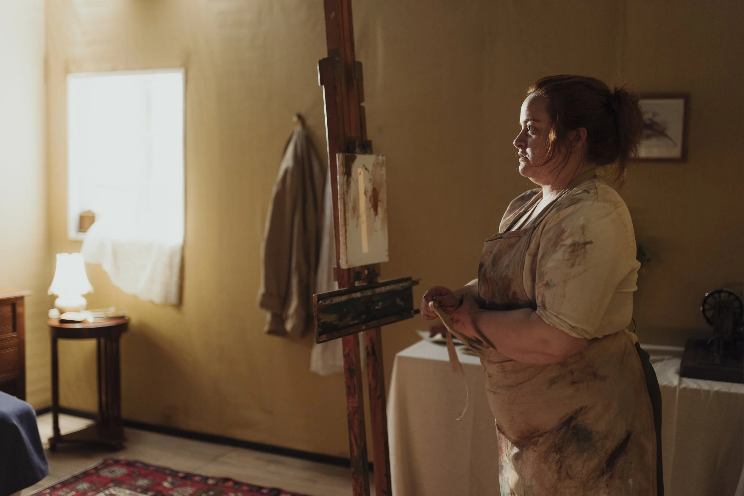 A woman standing in front of an easel painting in a warmly lit room with a window, a table with a cloth, and a framed picture on the wall.