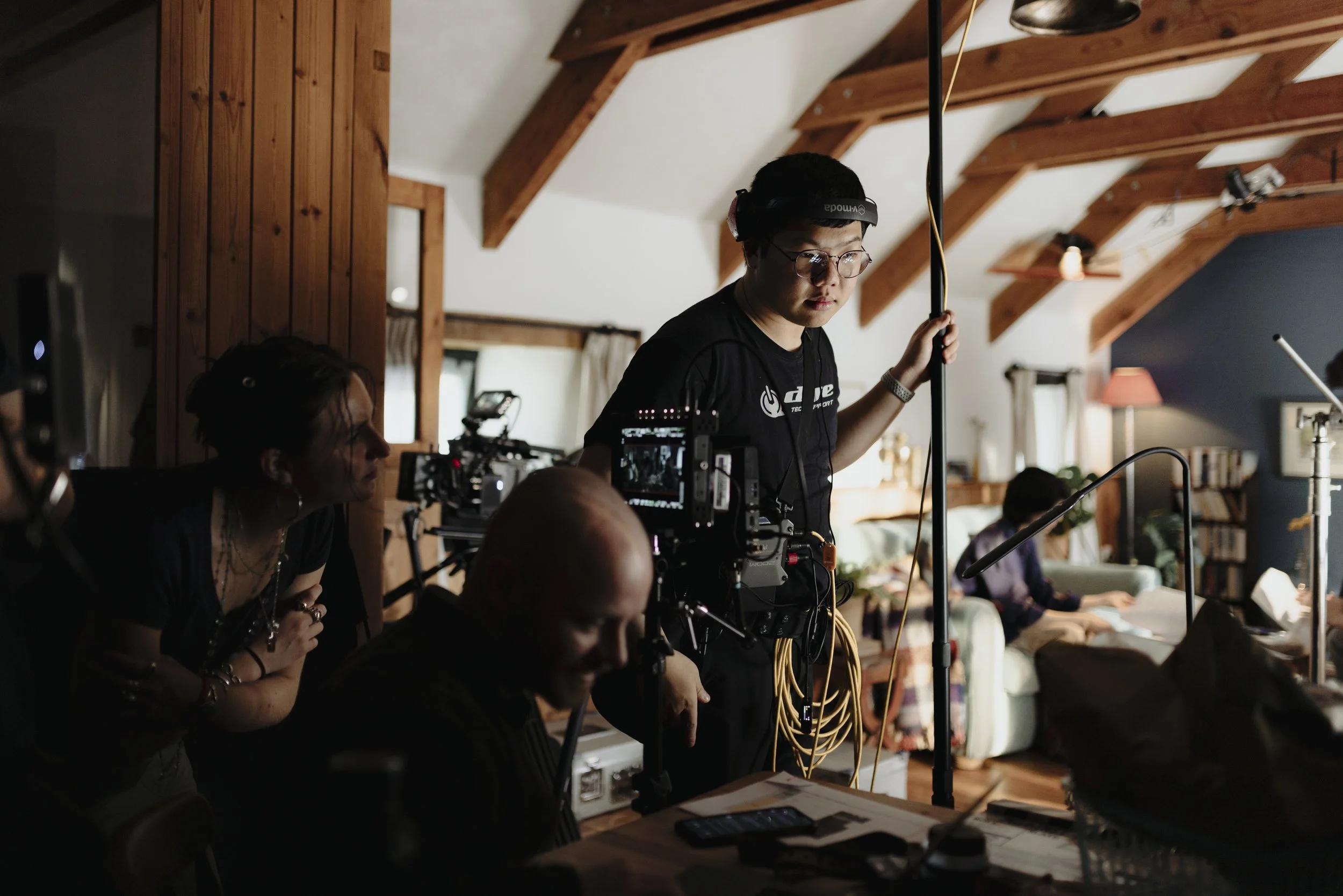 A film crew setting up equipment in a cozy living room with wooden beams on the ceiling, with several crew members focused on their tasks.