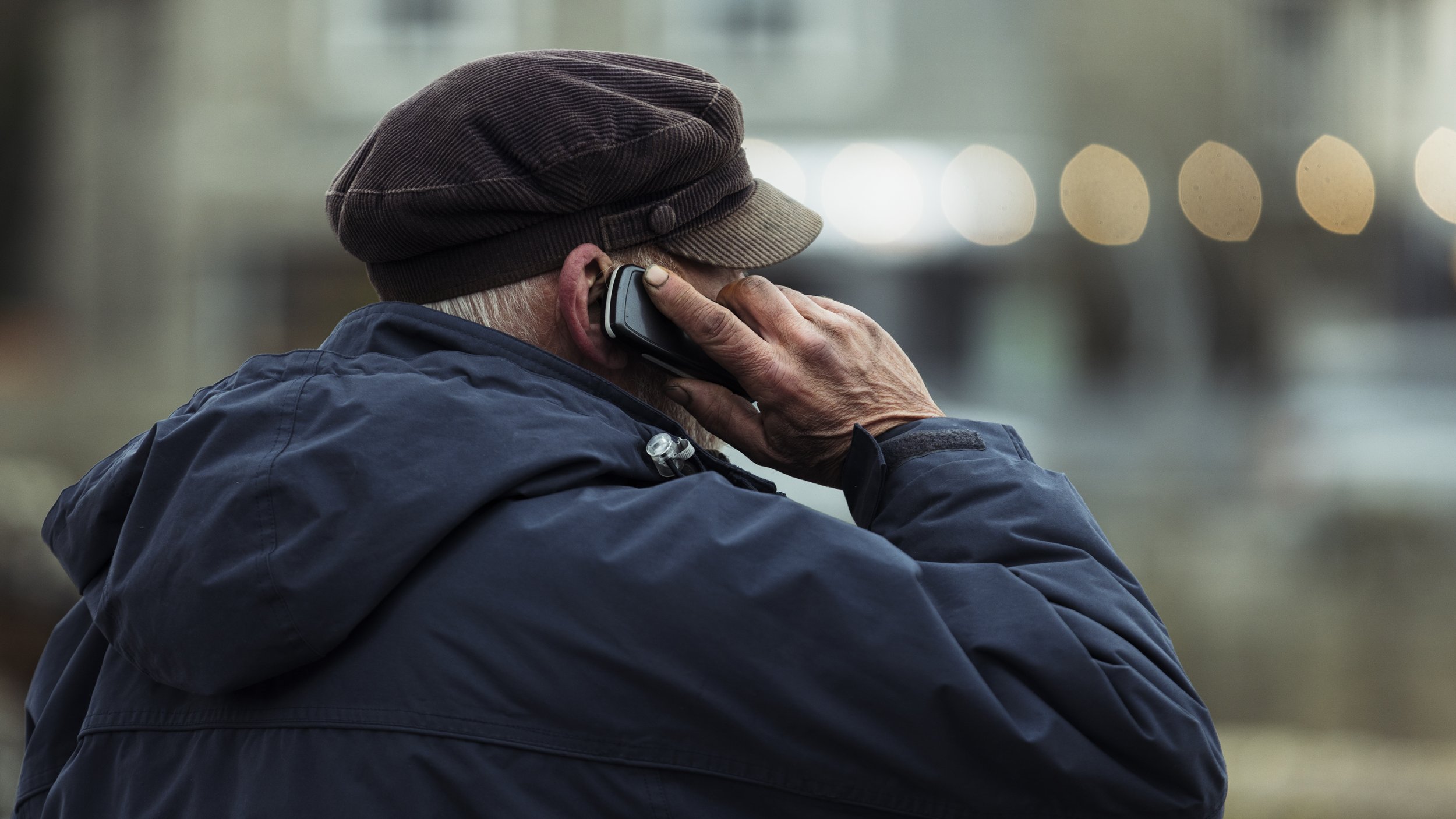 An elderly man wearing a brown newsboy cap and blue jacket talking on a cellphone with a blurred background.