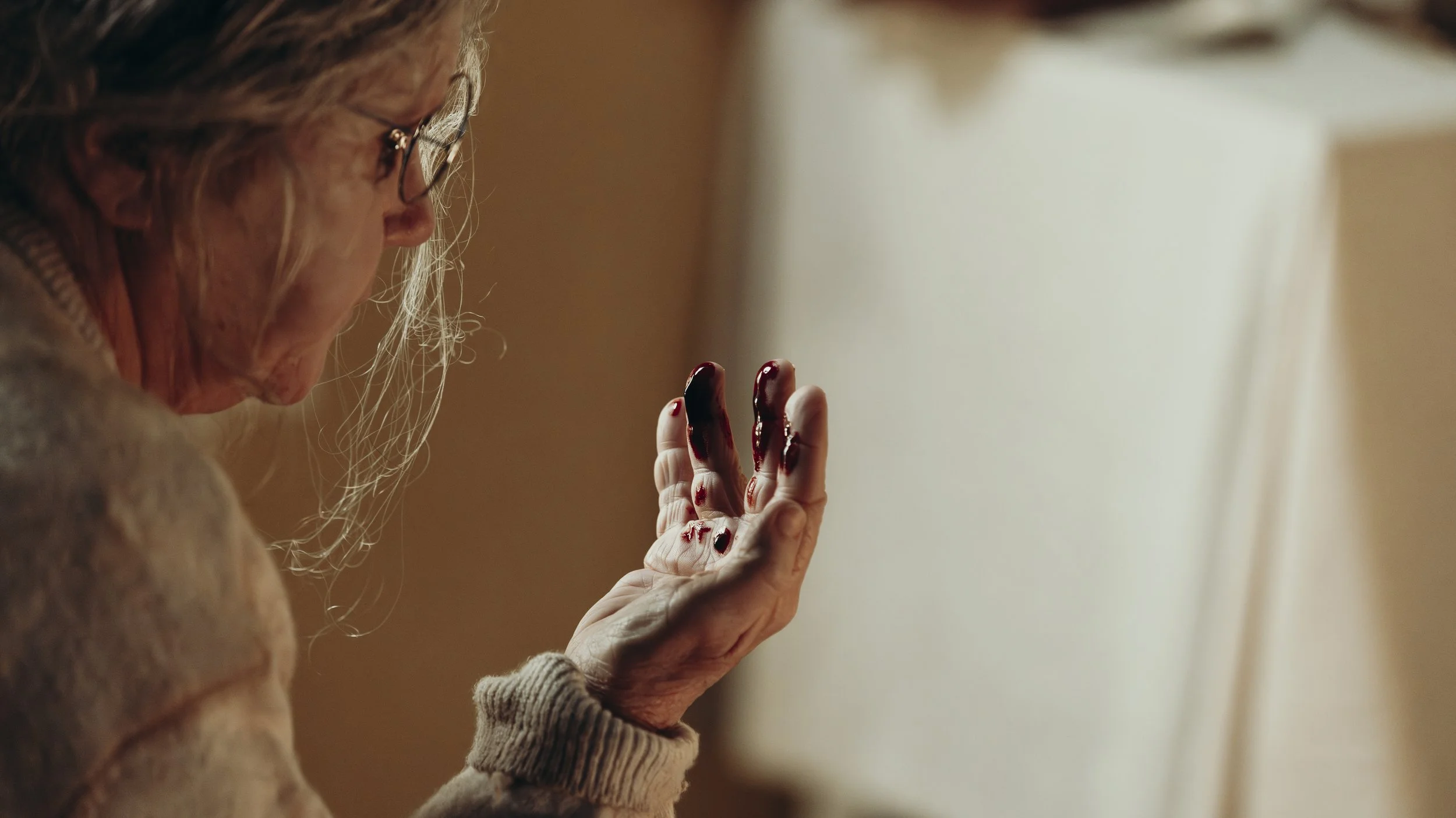 An elderly woman with glasses looks at her bloodstained hand, which is raised and has dark red blood on her fingers and palm.