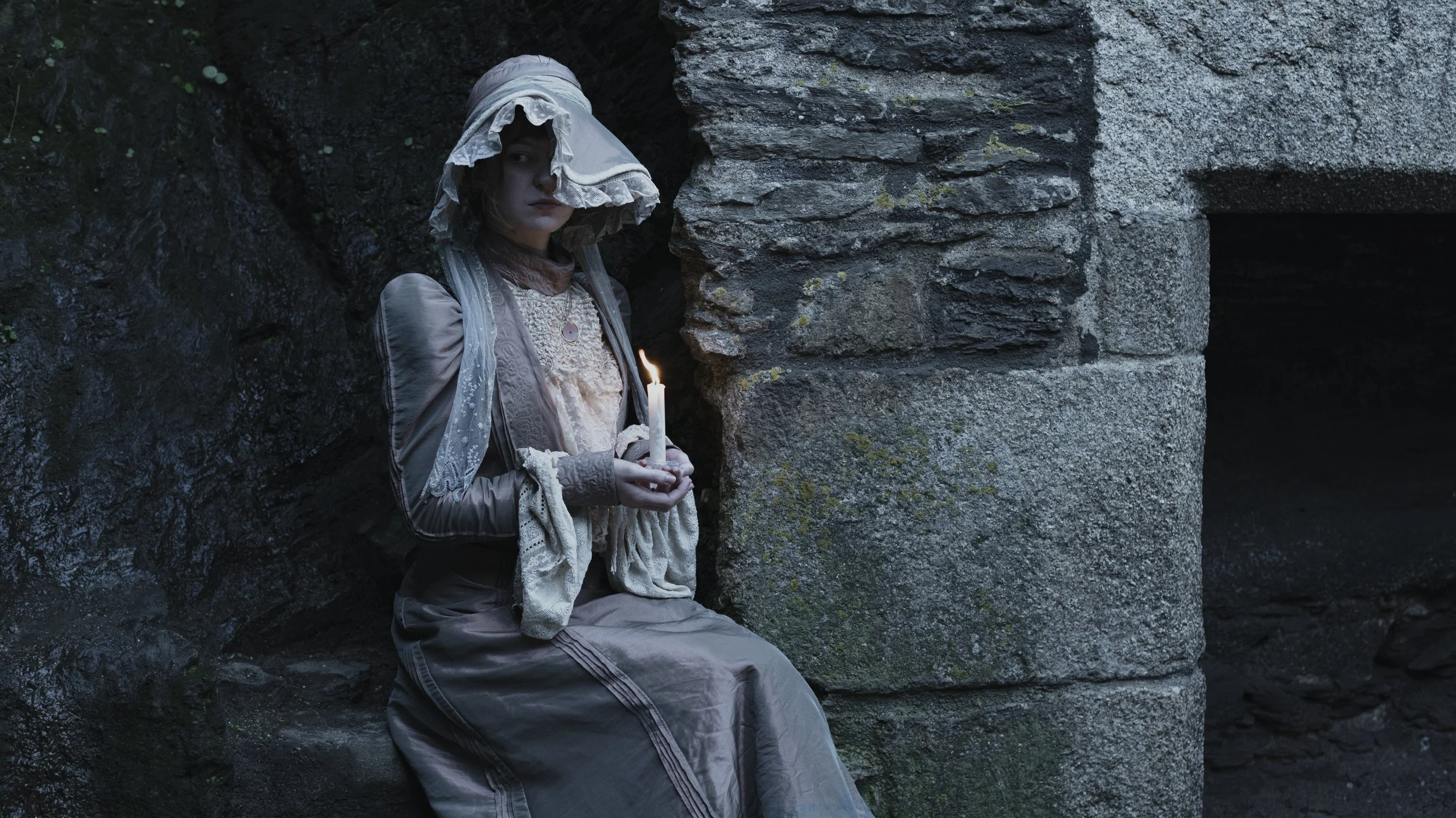 Woman dressed in vintage clothing, sitting against stone, holding a lit candle, wearing a bonnet, in a dark outdoor setting.