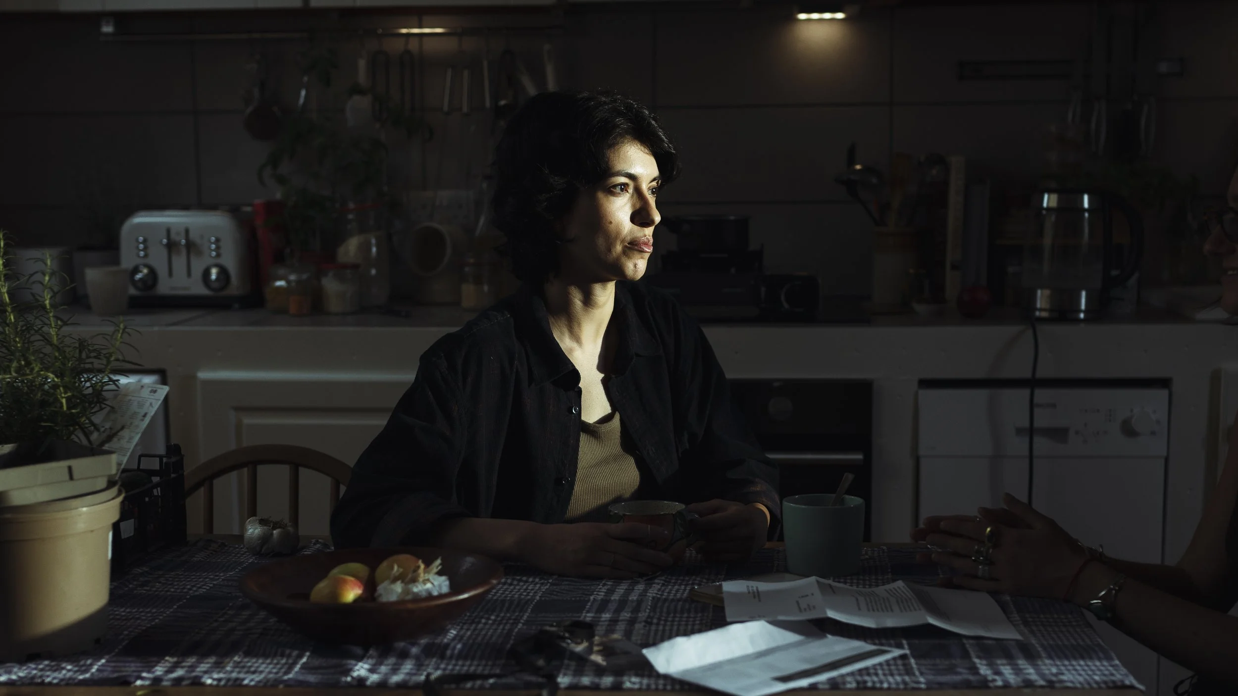 A woman with dark hair sitting at a kitchen table holding a cup, with natural light illuminating her face, and another person partially visible on the right side.
