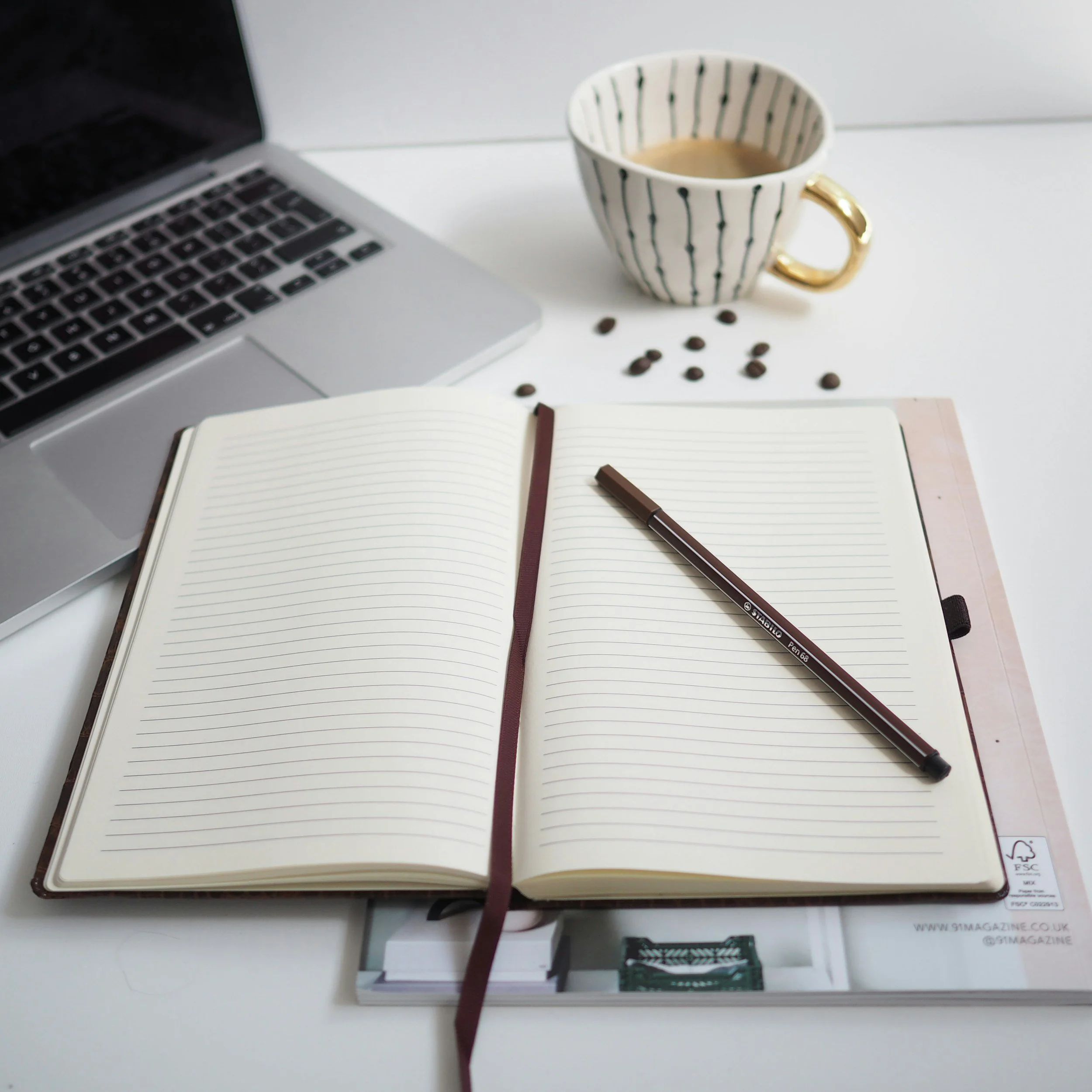 Open notebook with lined pages and a brown pen on top, a cup of coffee in a white mug with black stripes and a gold handle, scattered coffee beans, a silver laptop, and a magazine or brochure underneath the notebook on a white desk.