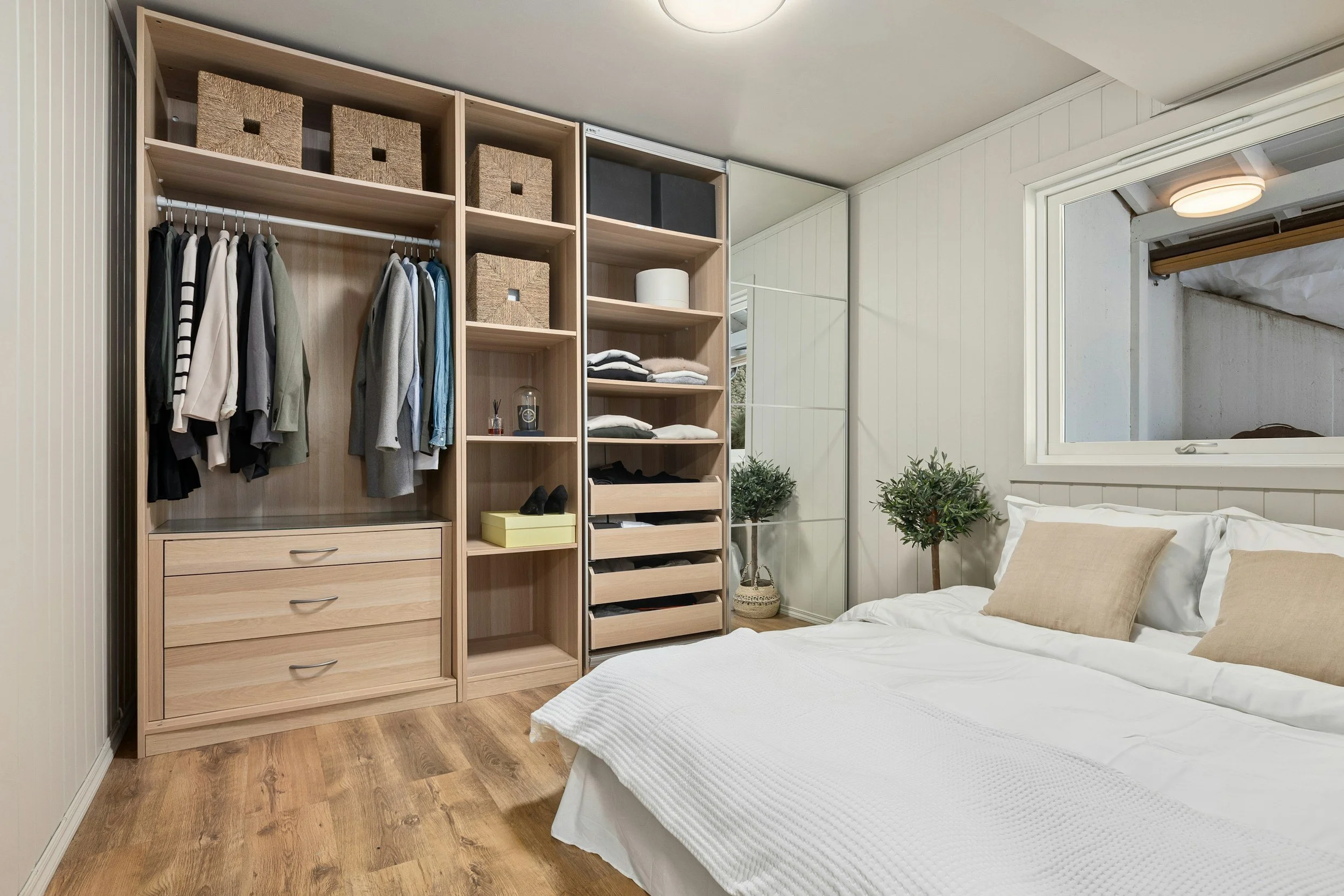 A bedroom with an open wooden closet containing hanging clothes, drawers, and storage boxes, next to a bed with beige pillows and white bedding, and a potted plant near a mirrored wardrobe.