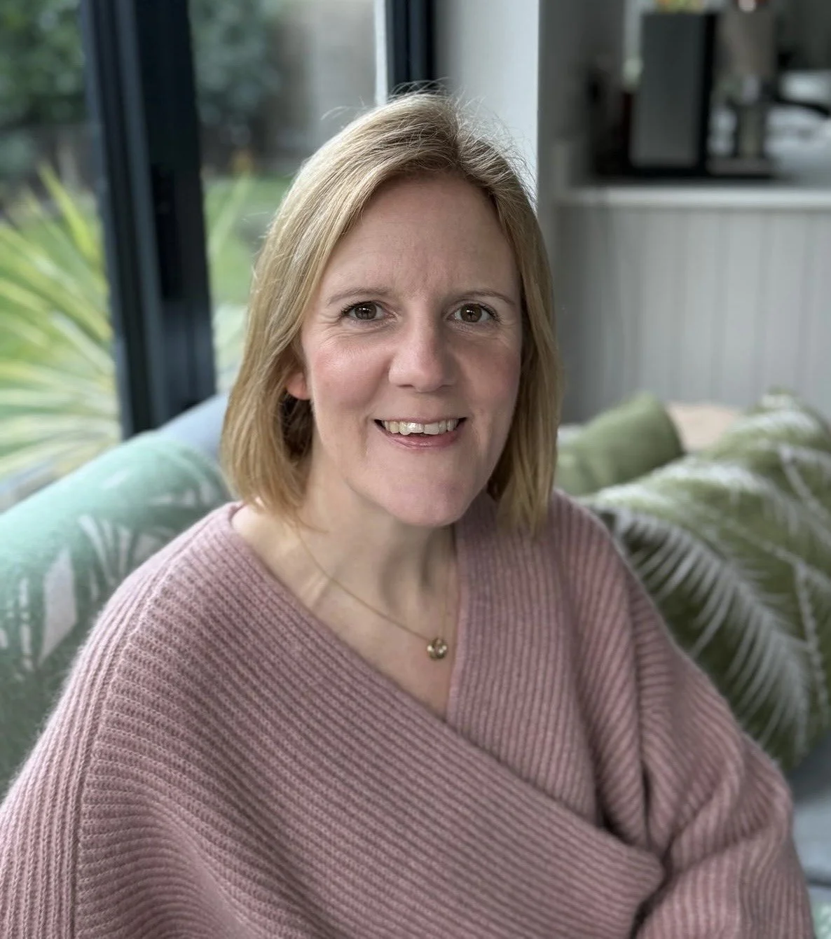A woman with blonde hair and a pink sweater sitting on a sofa in a well-lit room with large windows and green plants in the background.