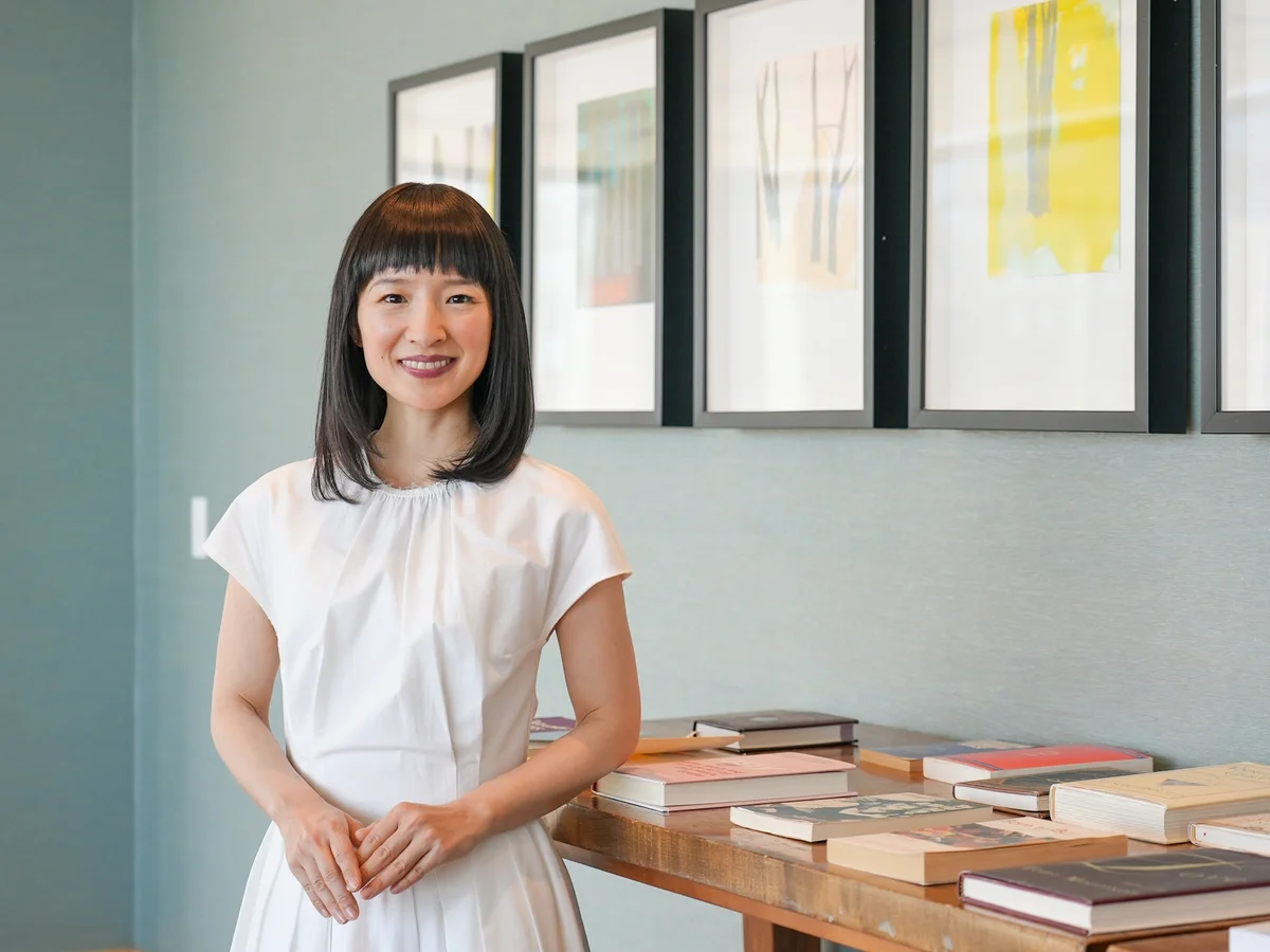 A woman with black hair and bangs, smiling, stands indoors near a table with several books. Behind her are framed pictures on a light-colored wall.