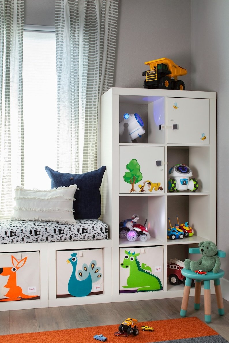 A child's playroom corner with a white storage unit filled with toys, including a robot and a construction vehicle, next to a window with patterned curtains. There are colorful animal-themed bins, a small green stuffed bear on a chair, and a few toy 