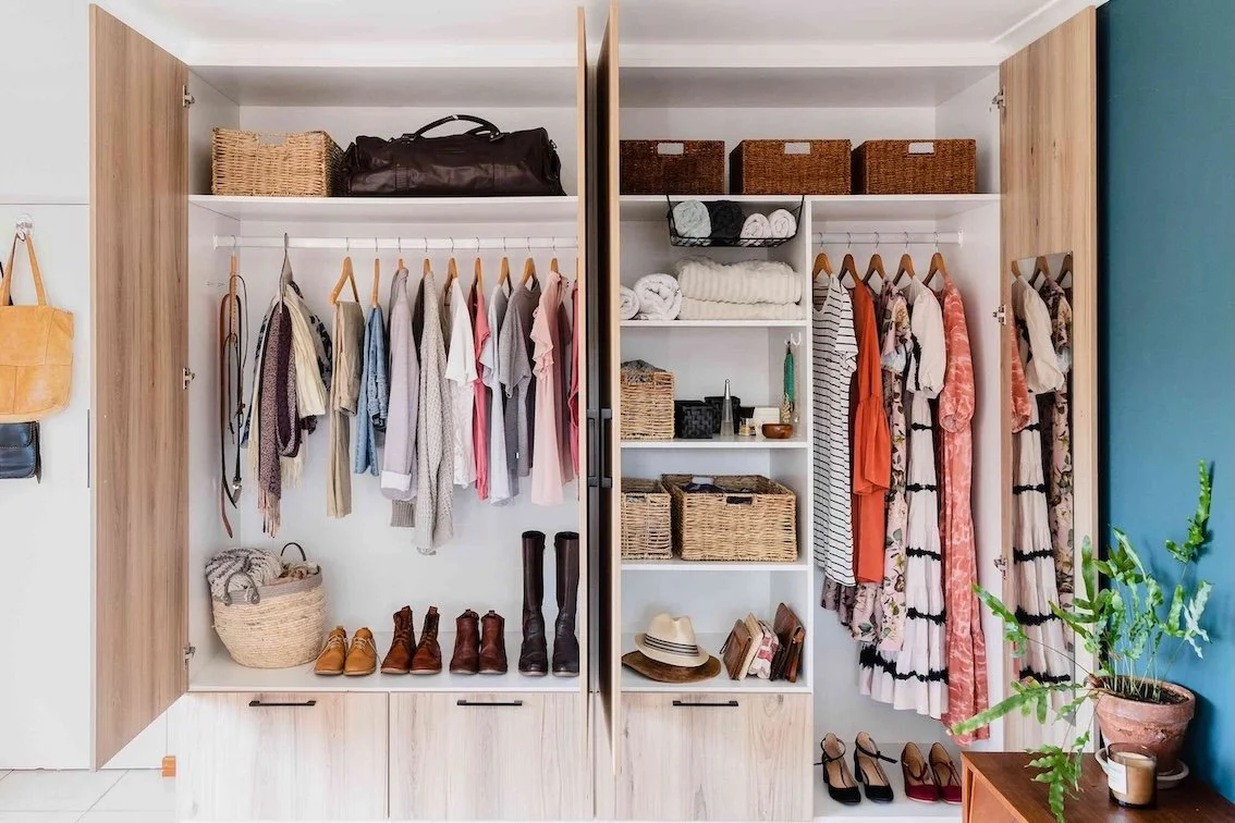 Open wardrobe with clothing, shoes, baskets, hats, and accessories, next to a blue wall and a potted plant.