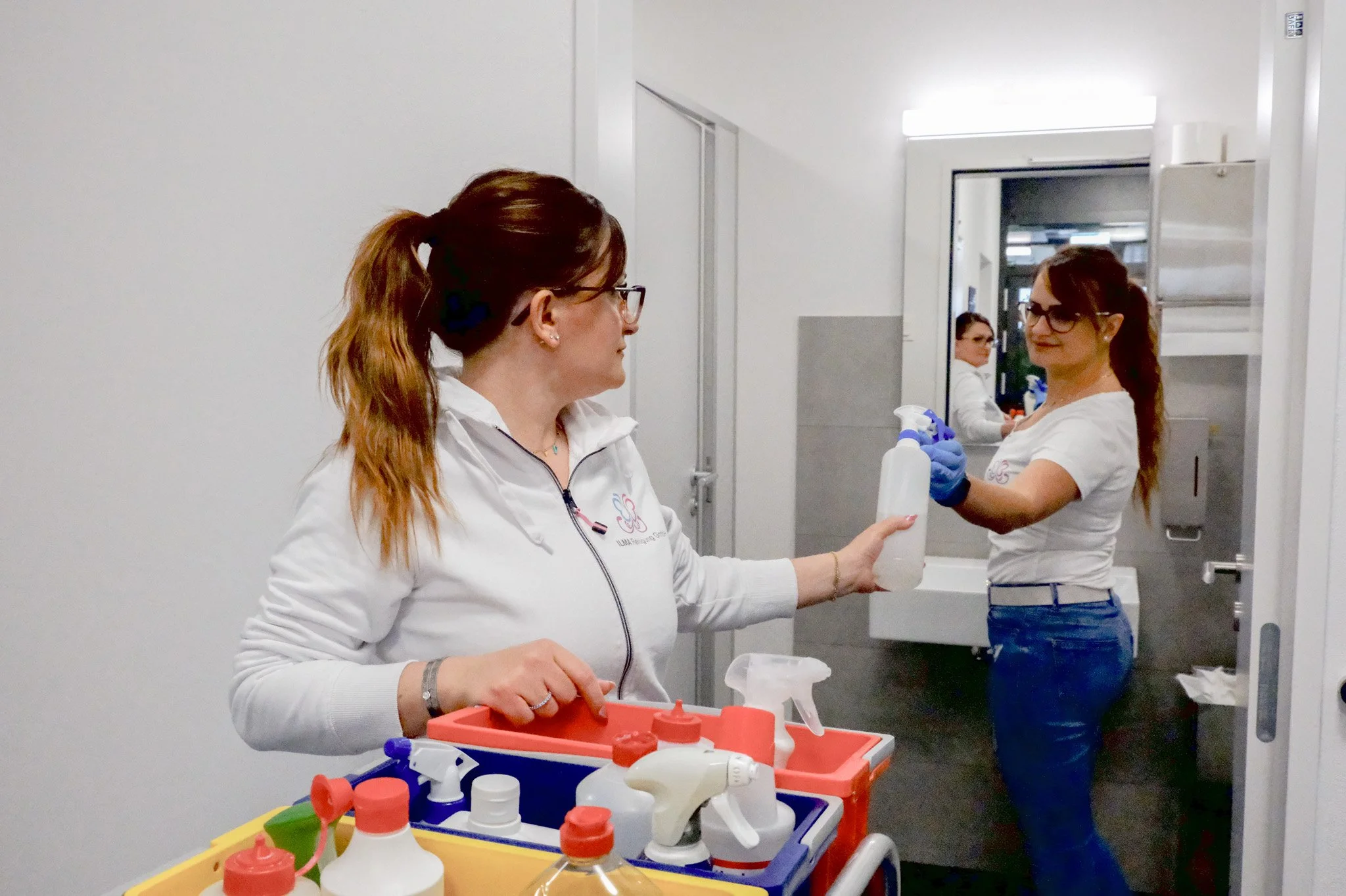 Zwei Frauen im Reinigungsraum mit Sprühflasche, Reinigungsmitteln und Utensilien auf einem Wagen.