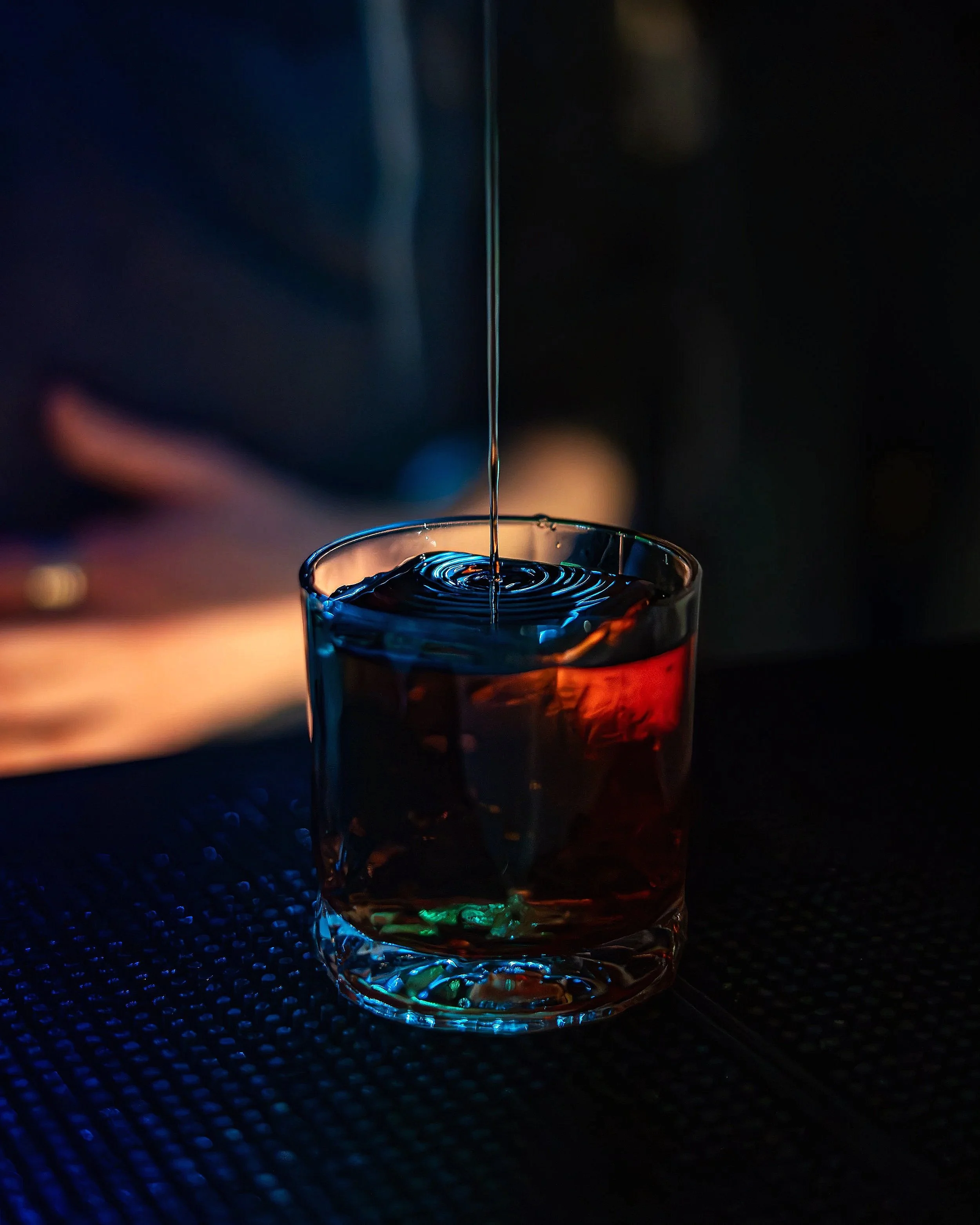 A glass with a dark-colored beverage, possibly whiskey or rum, sitting on a textured dark surface. A stream of clear liquid is being poured into the glass in a dimly lit setting.