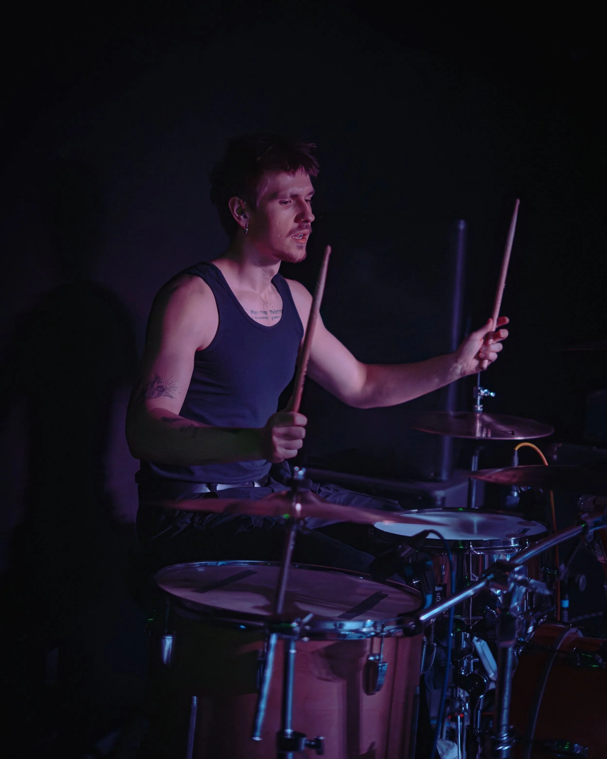 A drummer playing on a drum set in a dark, possibly concert setting.