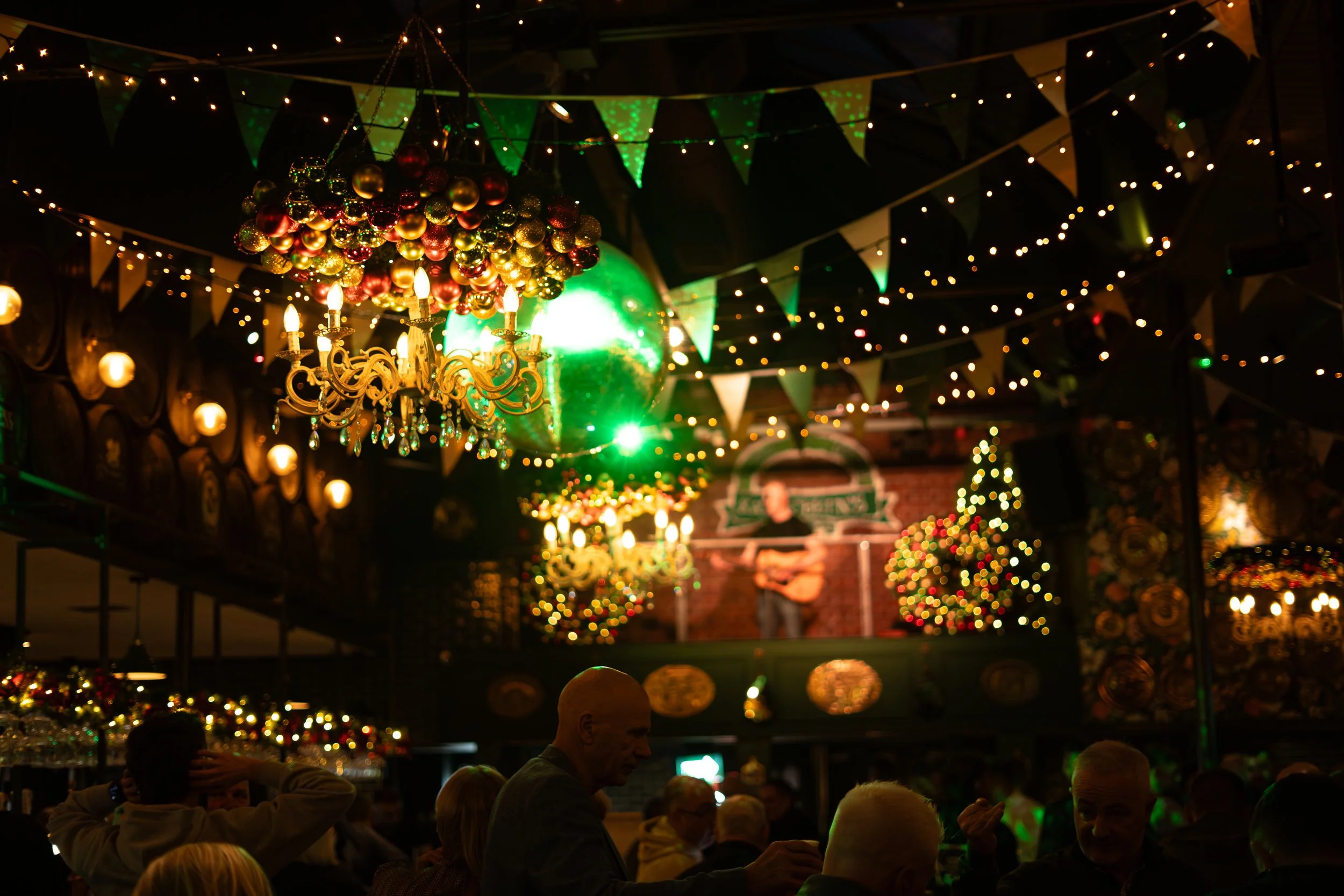 People attending a festive event in a decorated indoor venue with Christmas trees and string lights, featuring a performer playing guitar on stage.