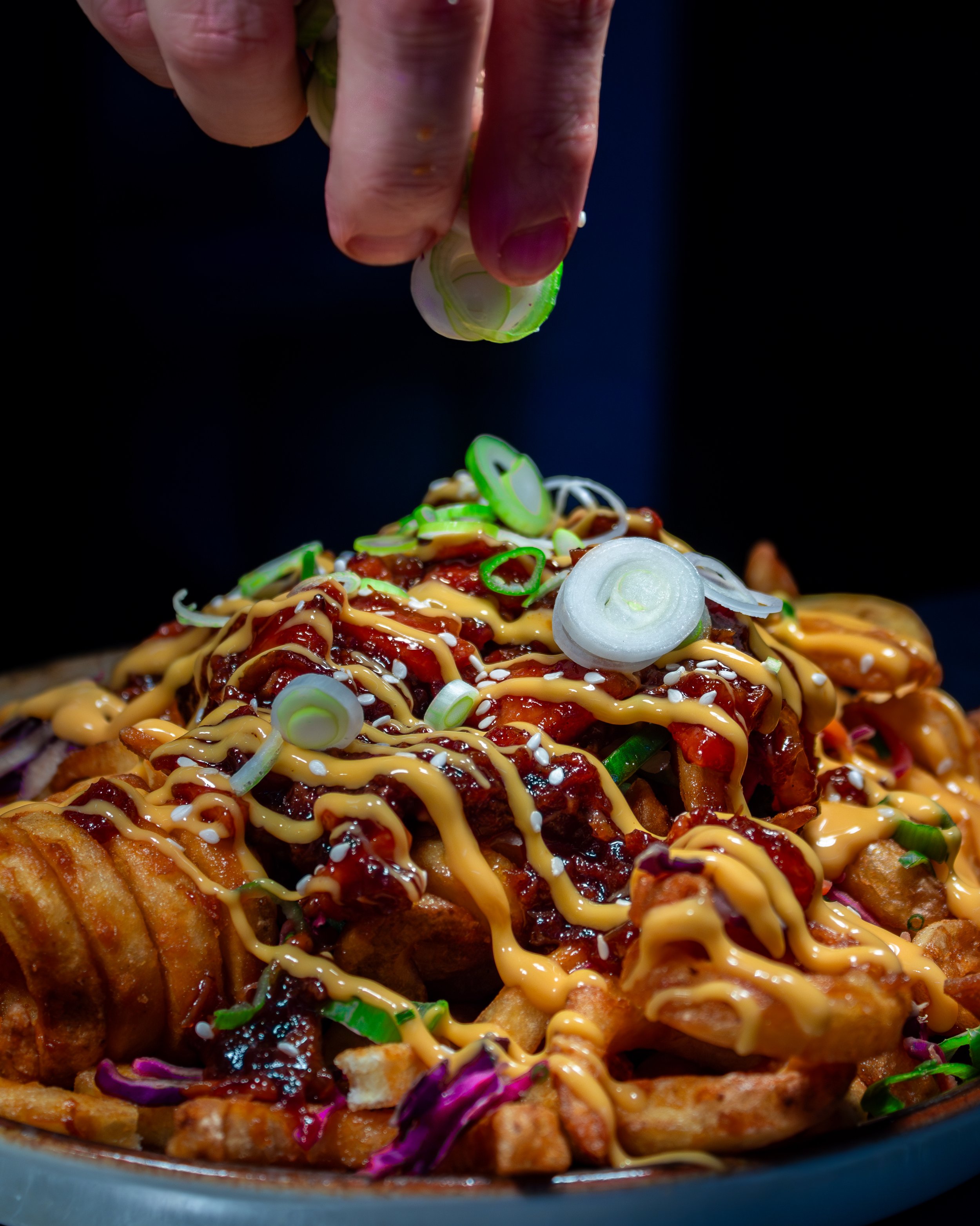 A hand sprinkling chopped green onions on a plate of loaded fries.
