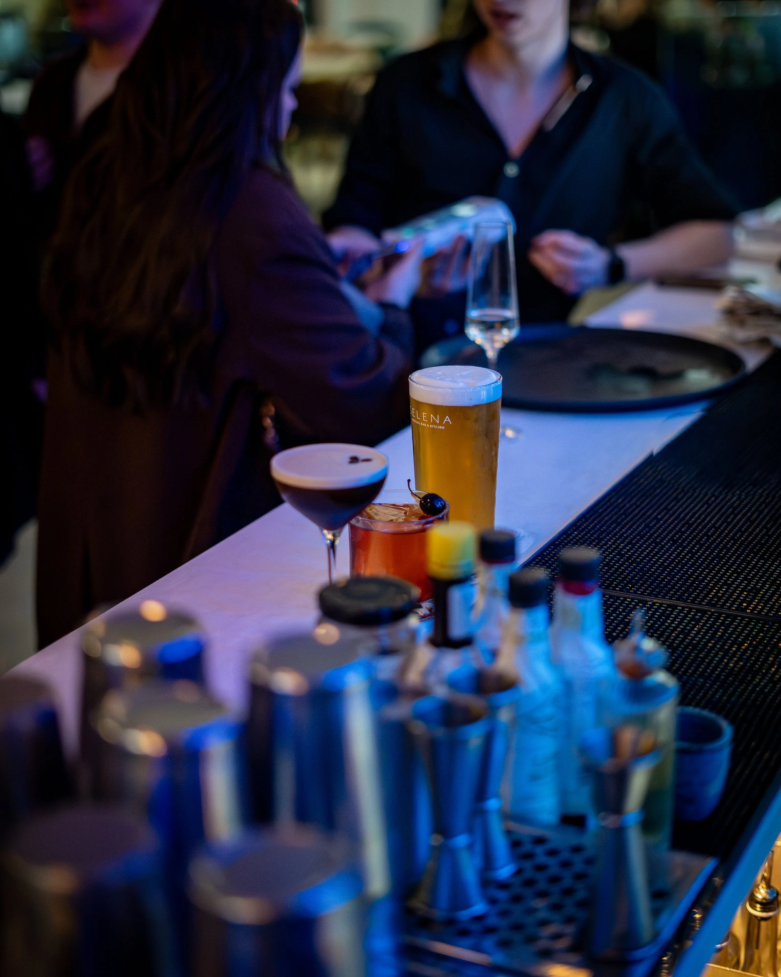 Bar countertop with various cocktails and beer, two women talking in the background in a dimly lit environment.