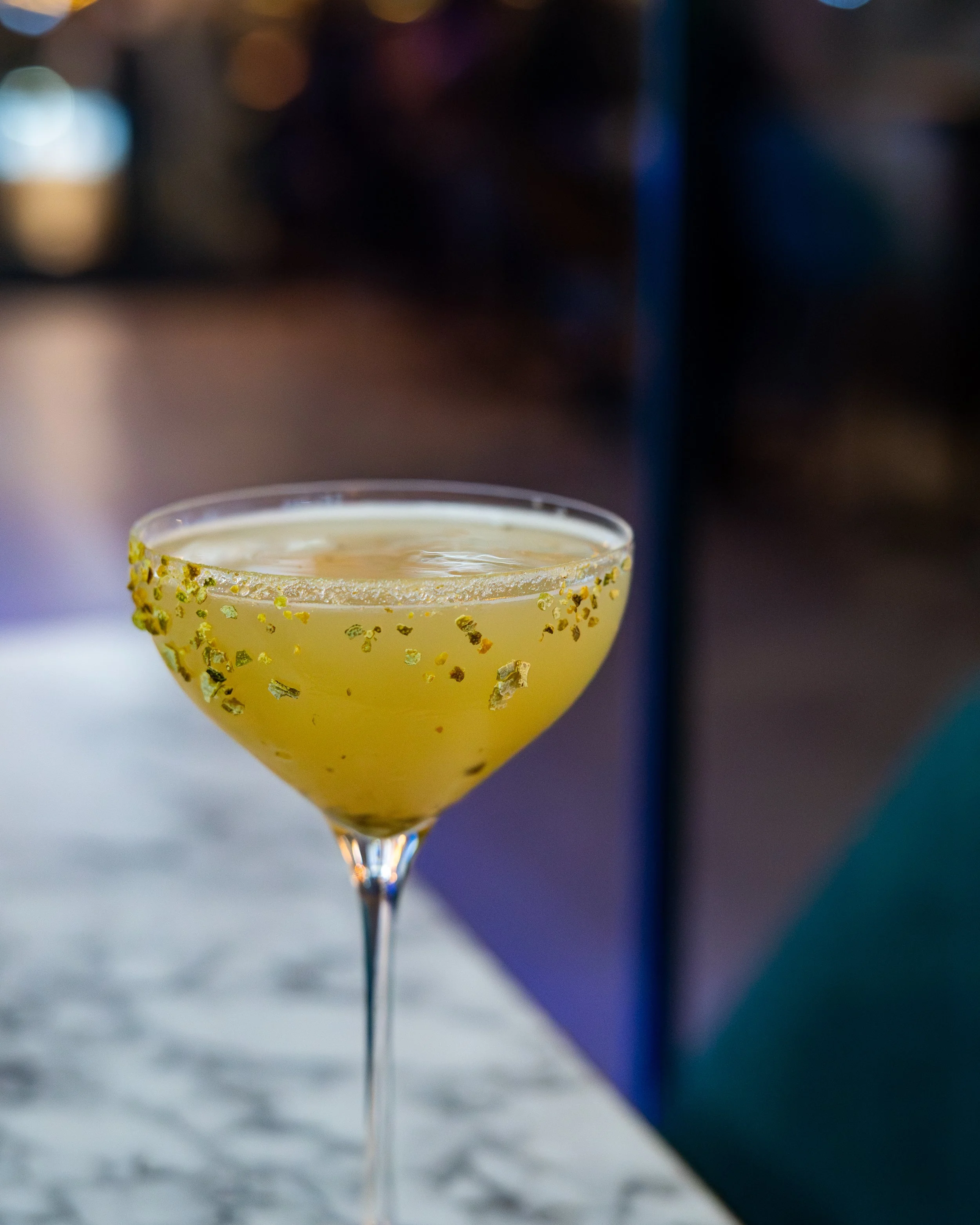 Cocktail glass with a yellow-green drink, rimmed with gold-colored glitter, on a marble surface with a blurred background.