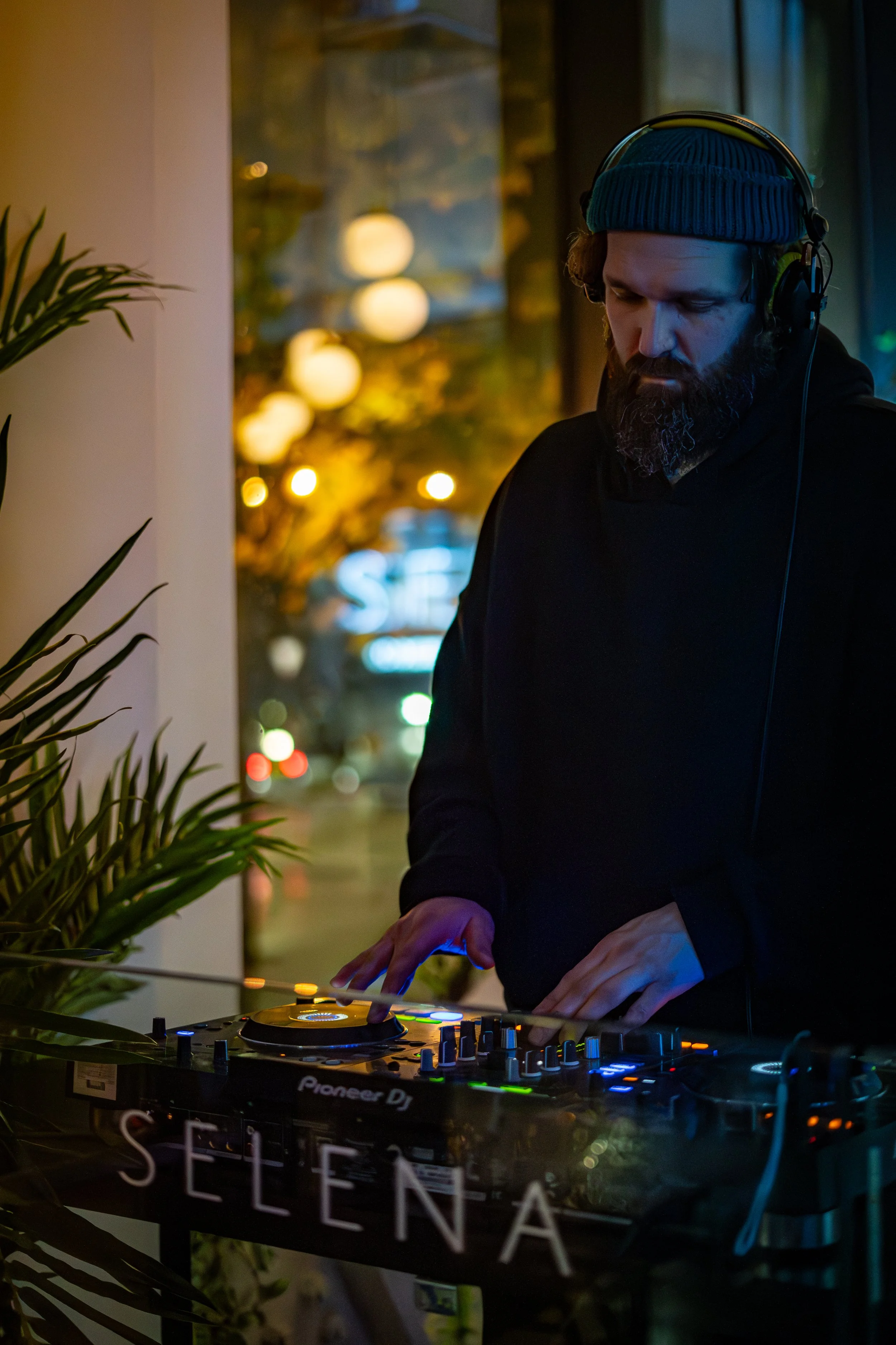A bearded man wearing a blue beanie and black hoodie DJing with headphones on in a dimly lit venue, with a mixer and turntable in front of him and alight plants nearby.