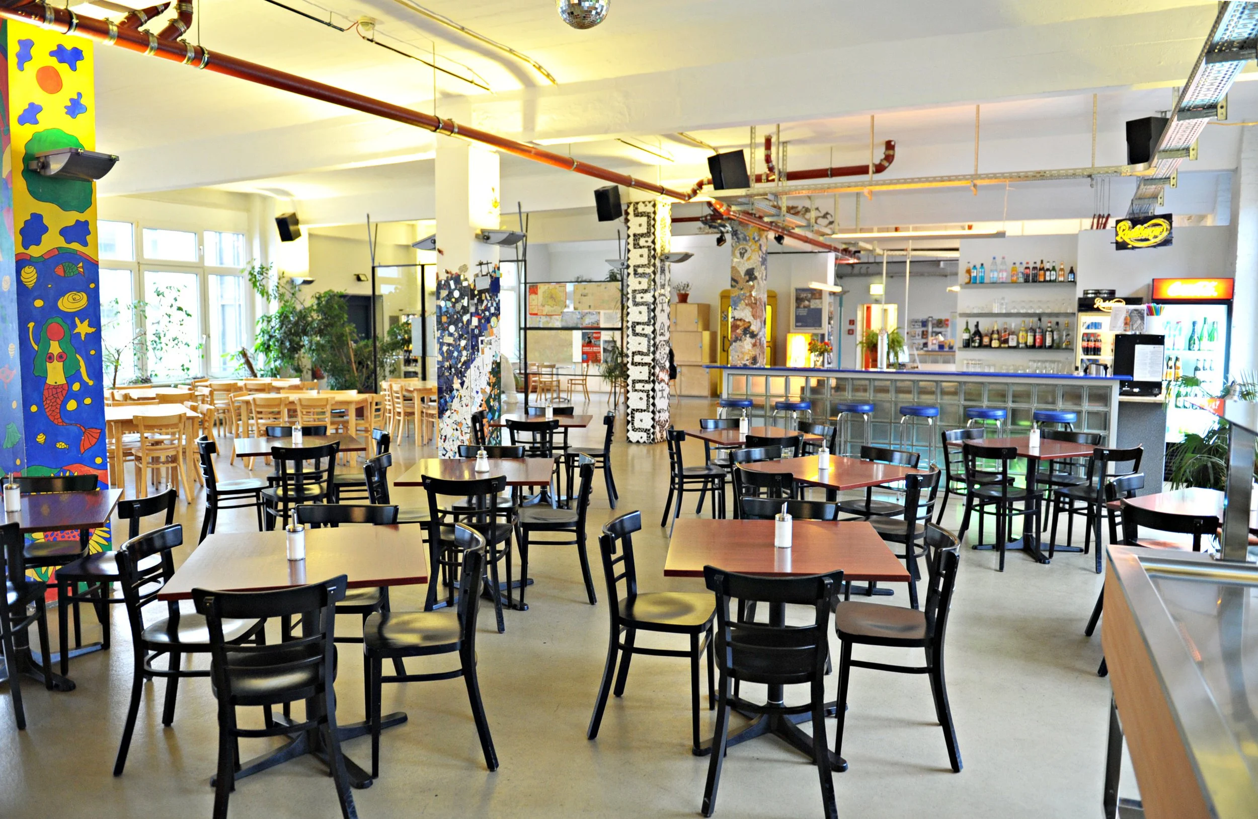 Der Frühstücksraum des Hotel Transit Loft mit Tischen und Stühlen, bunter Wanddekoration, Bar mit Getränken, Pflanzen und große Fenster, viel natürliches Licht.