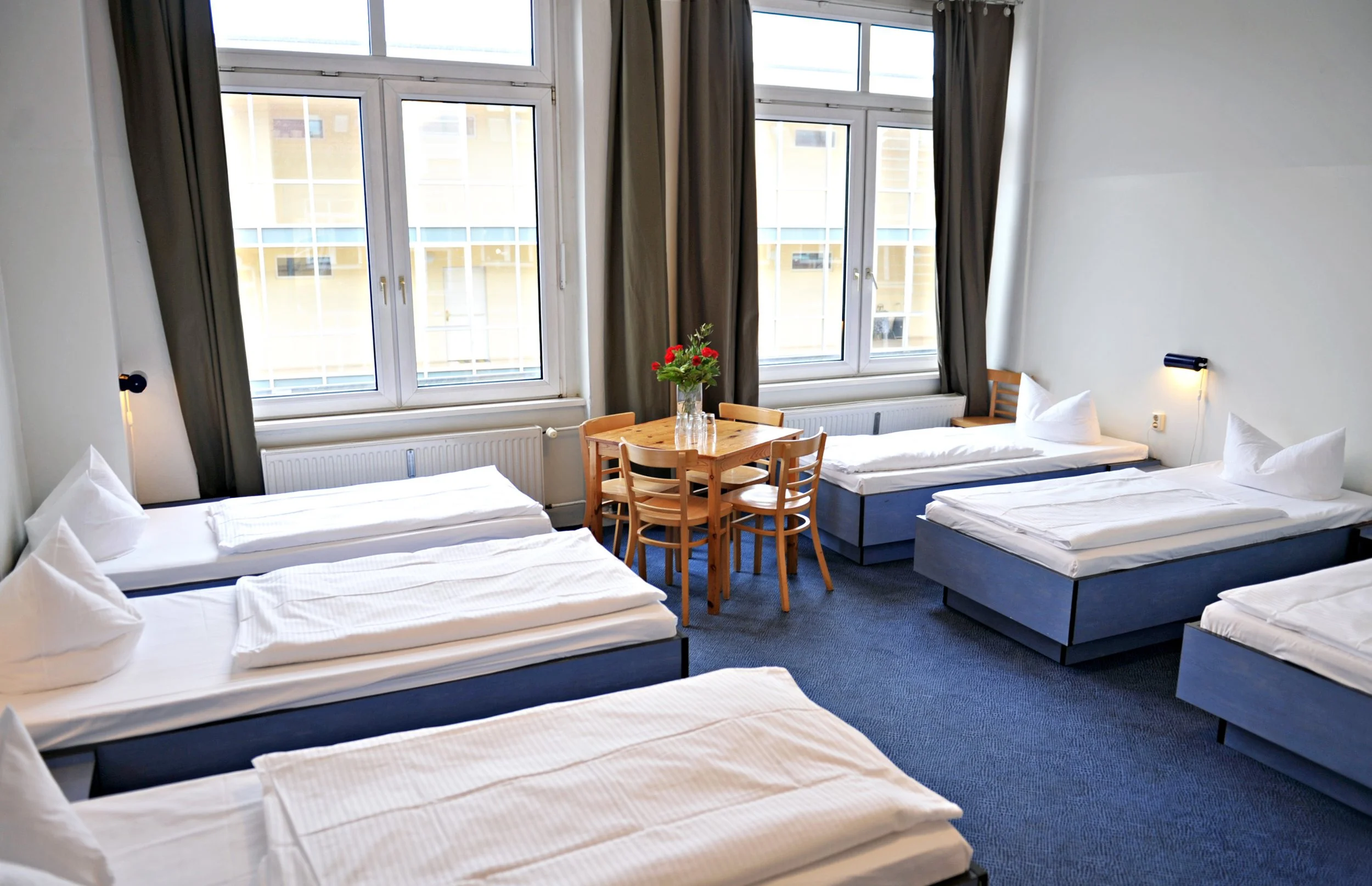 Ein Zimmer des Hotel Transit Loft mit vier Betten, einem runden Holztisch mit vier Stühlen und einem Tisch mit Blumenvase, große Fenster mit dunklen Vorhängen, weiß gestrichene Wände, blaue Teppichböden, Wandlampe über den Betten.