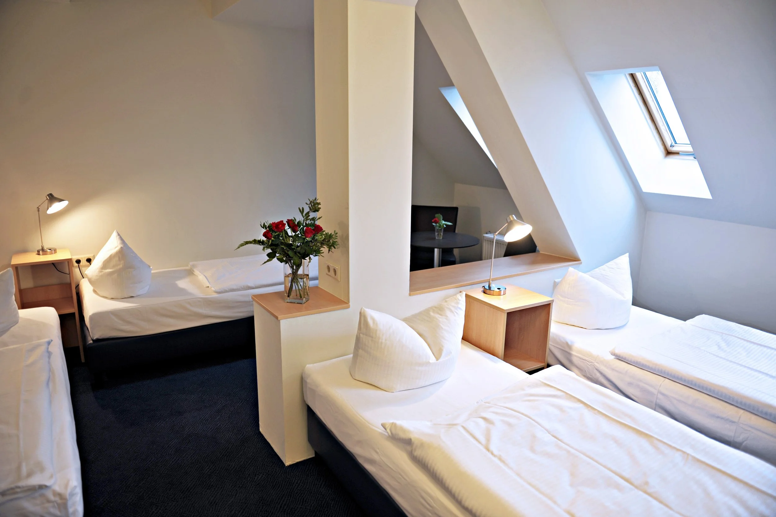 Ein Hotelzimmer mit drei Betten, einem kleinen Tisch mit Vase und Blumen, Leselampen, Dachfenstern und weißen Wänden.