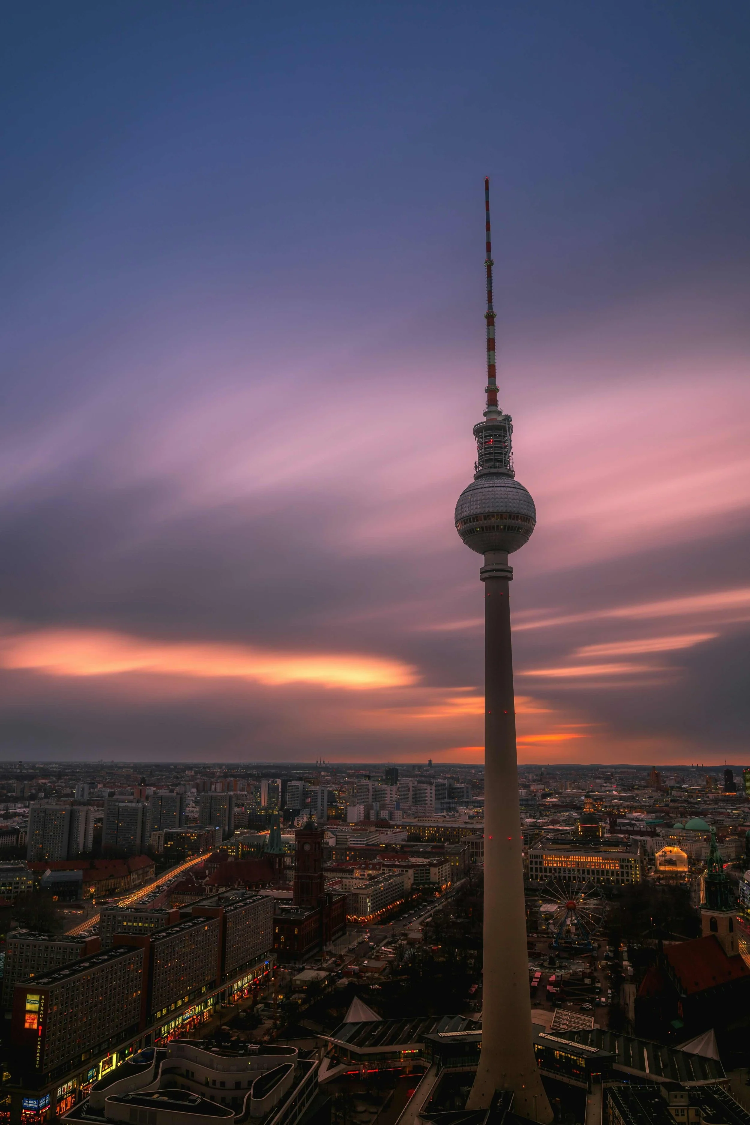 Blick auf den Berliner Fernsehturm während des Sonnenuntergangs, mit Wolken und einer Stadtansicht im Vordergrund.