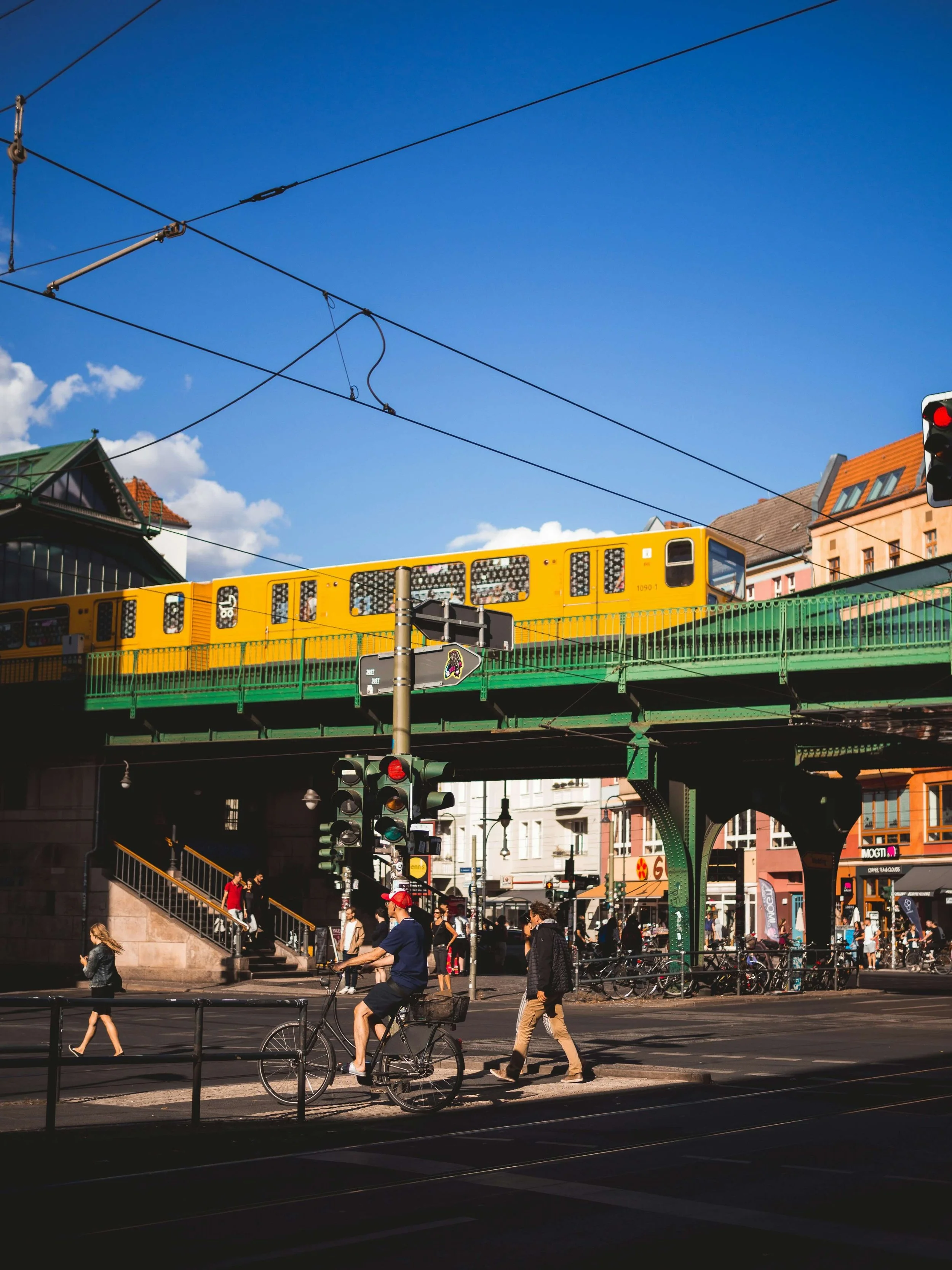 Stadtbild mit Bahnbrücke, gelbem Zug und Fußgängern, Fahrradfahrern bei sonnigem Himmel.