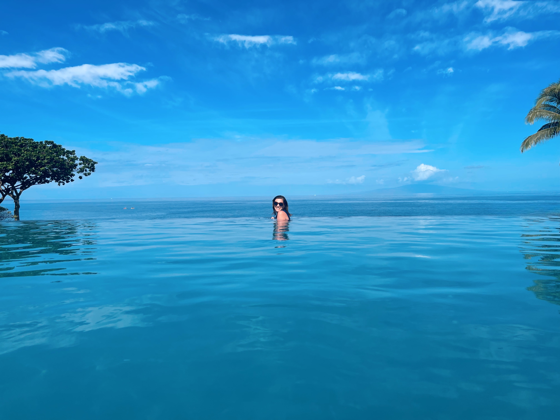 A woman in sunglasses swimming in an infinity pool overlooking the ocean under a blue sky with scattered clouds, with trees on either side.