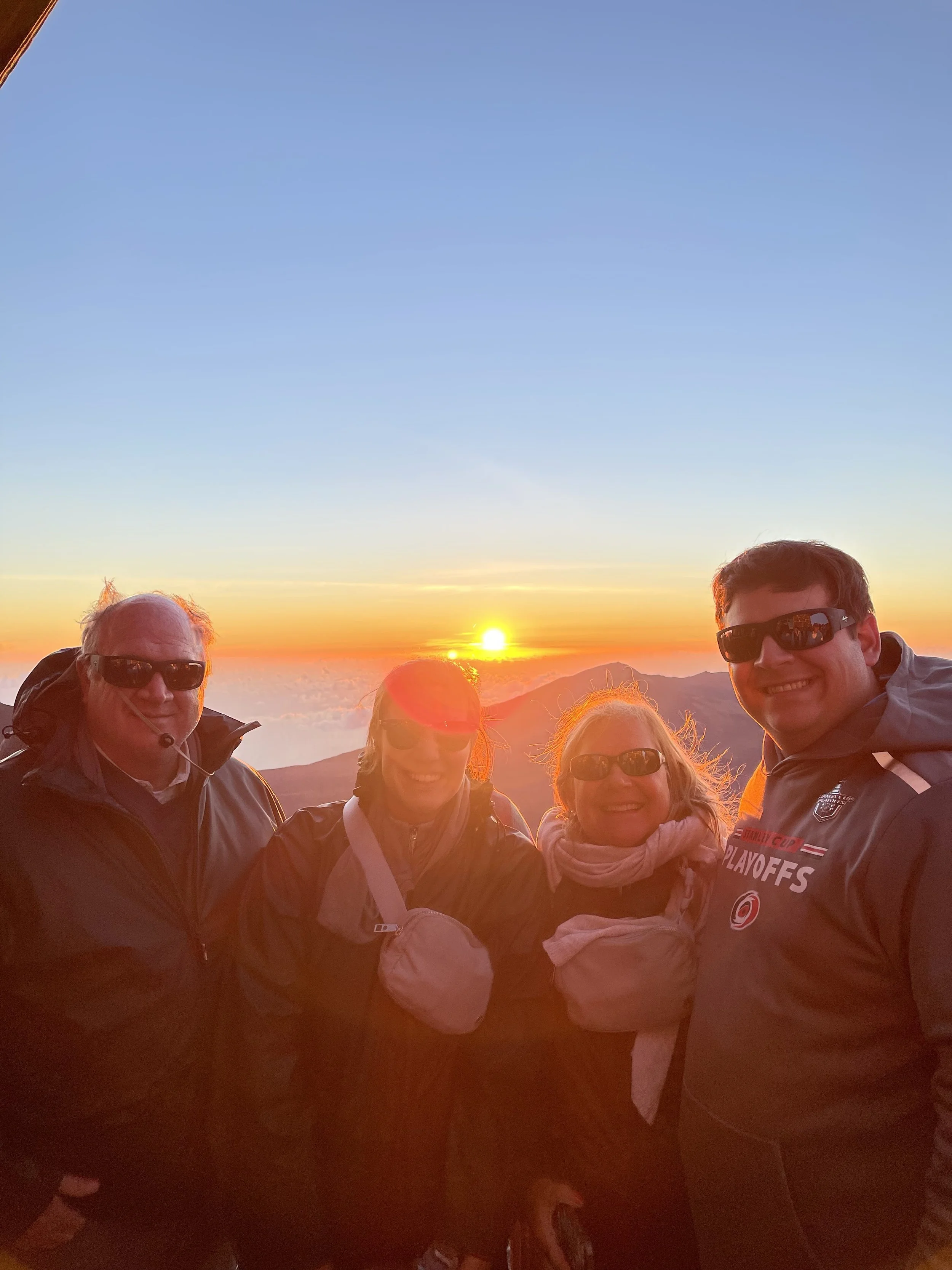 Group of four people standing on a mountain at sunrise, wearing jackets and sunglasses, with mountains and a partly cloudy sky in the background.