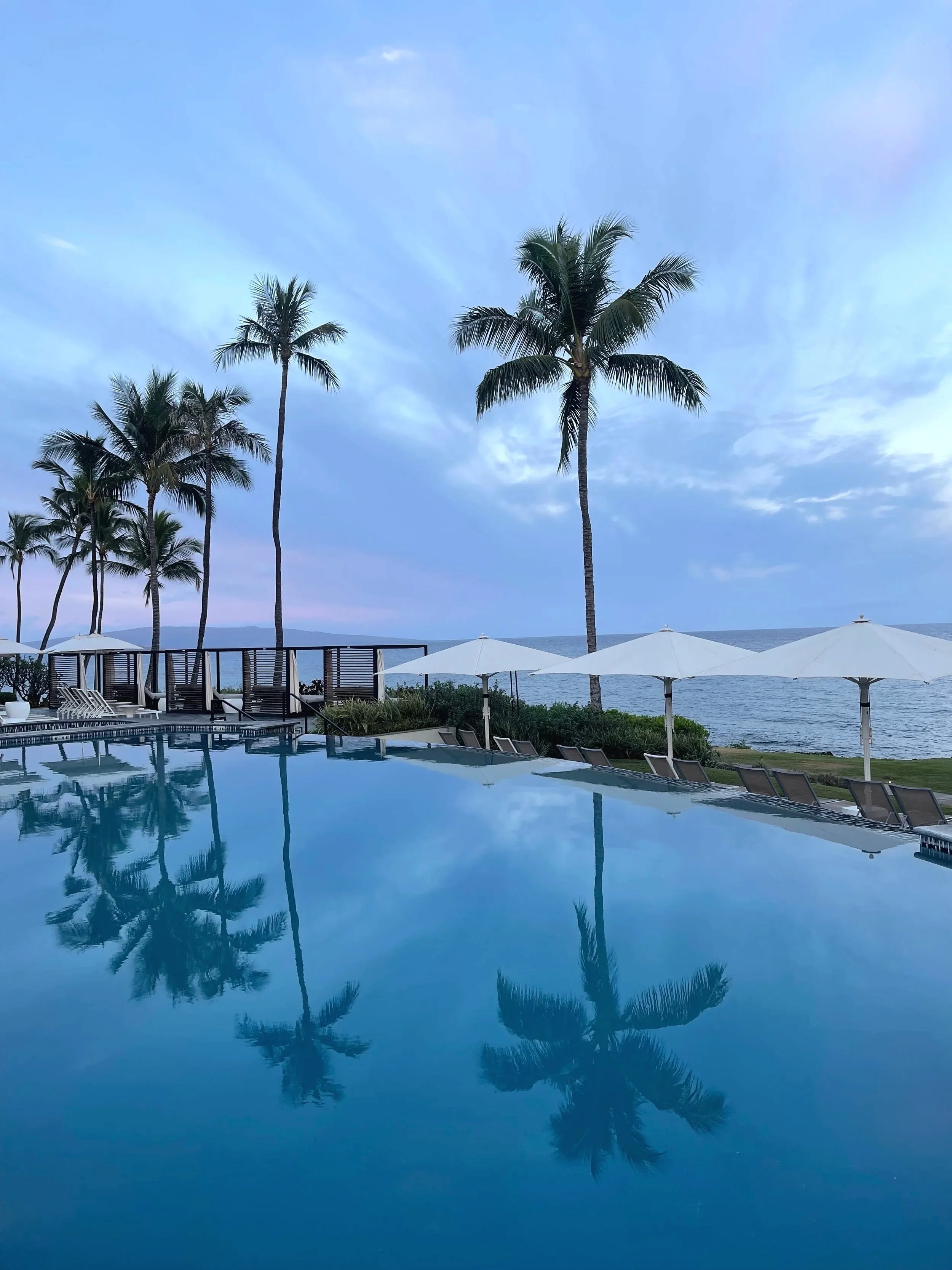 Swimming pool with lounge chairs and white umbrellas, palm trees, ocean view, and cloudy sky.