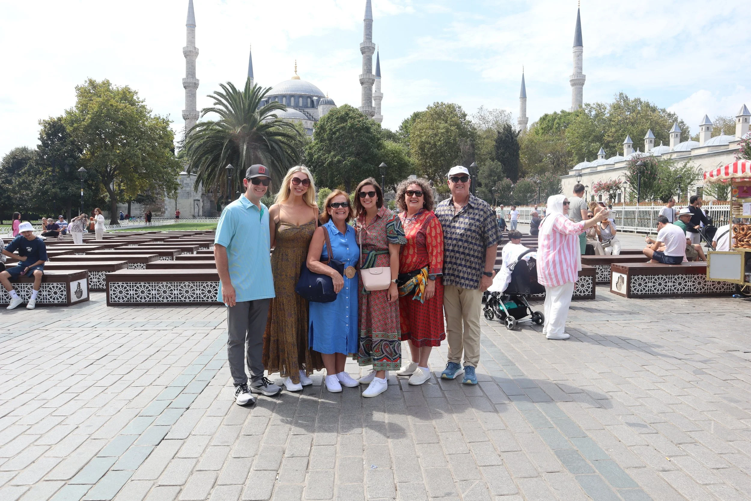 Group of six people smiling and posing for a photo in front of a historic mosque with multiple minarets, surrounded by trees and people sitting on benches and walking in a crowded outdoor area.