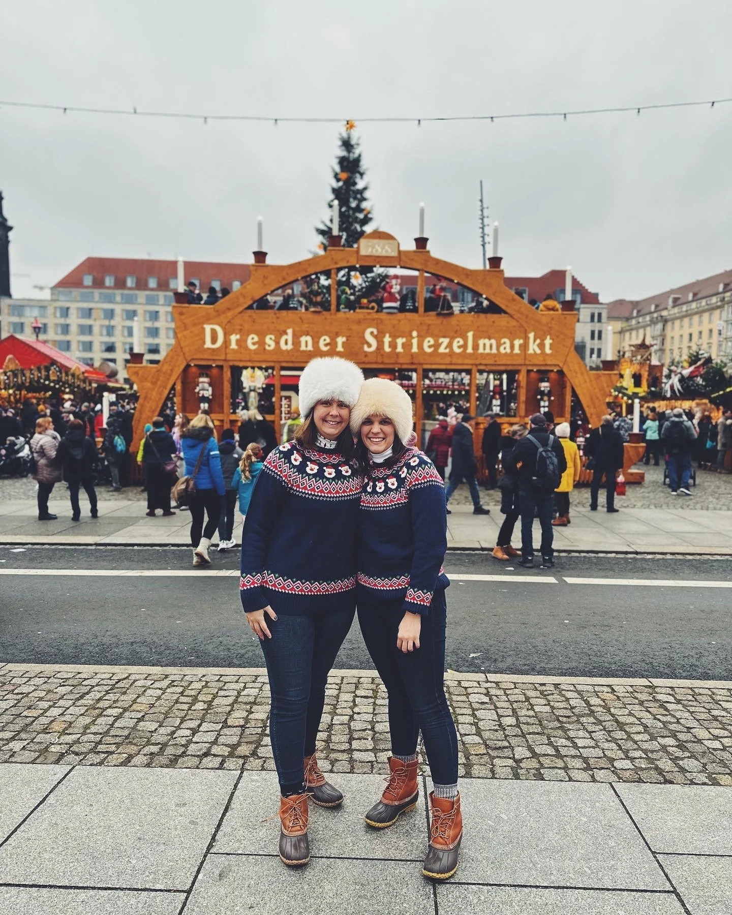 Two women dressed in matching Christmas sweaters and furry hats standing and smiling in front of a decorated Christmas market stall with a sign that reads 'Dresdner Striezelmarkt'.