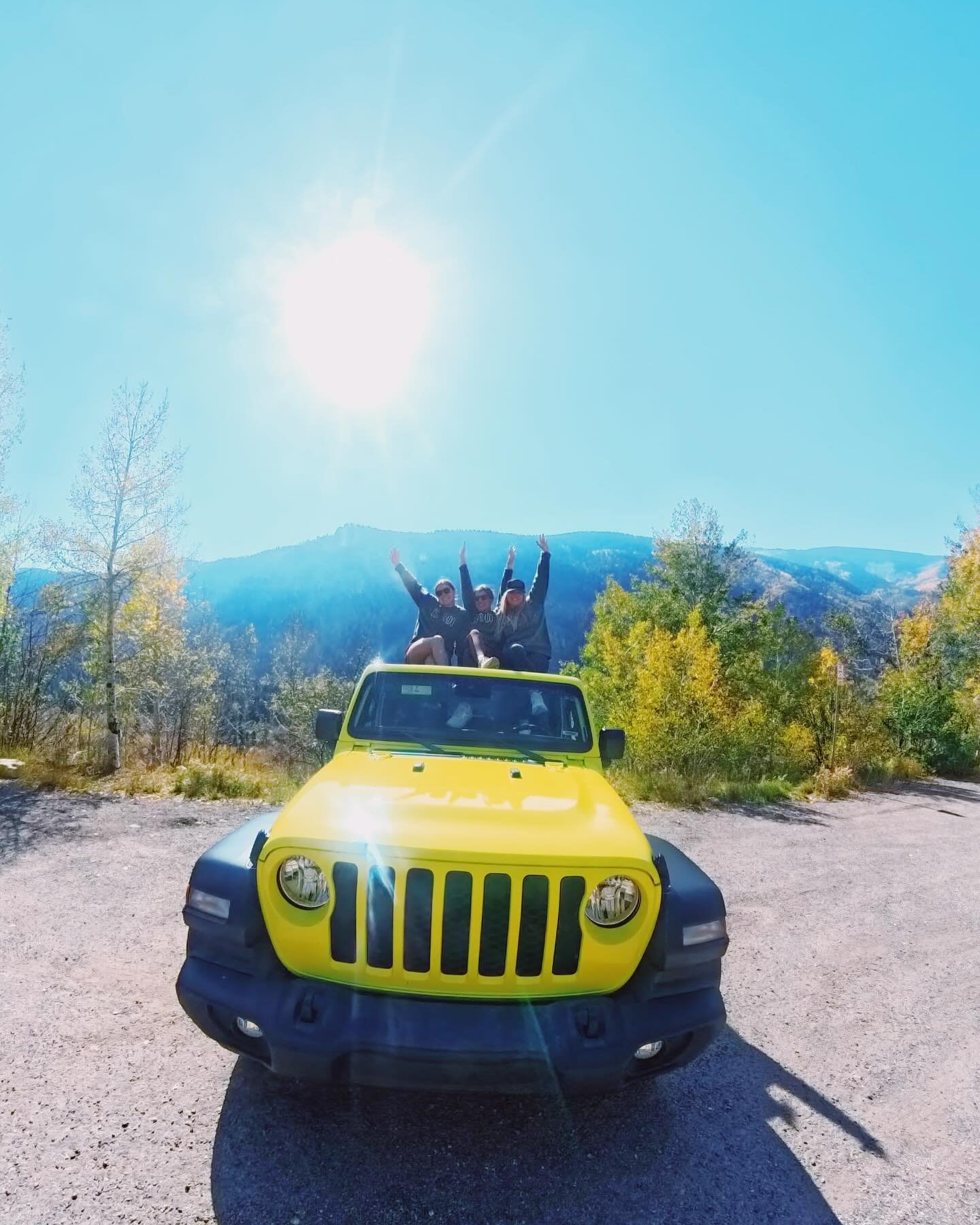 Three people sitting on top of a yellow Jeep with black wheel fenders, in a scenic outdoor area with trees and mountains in the background, under bright sunlight.