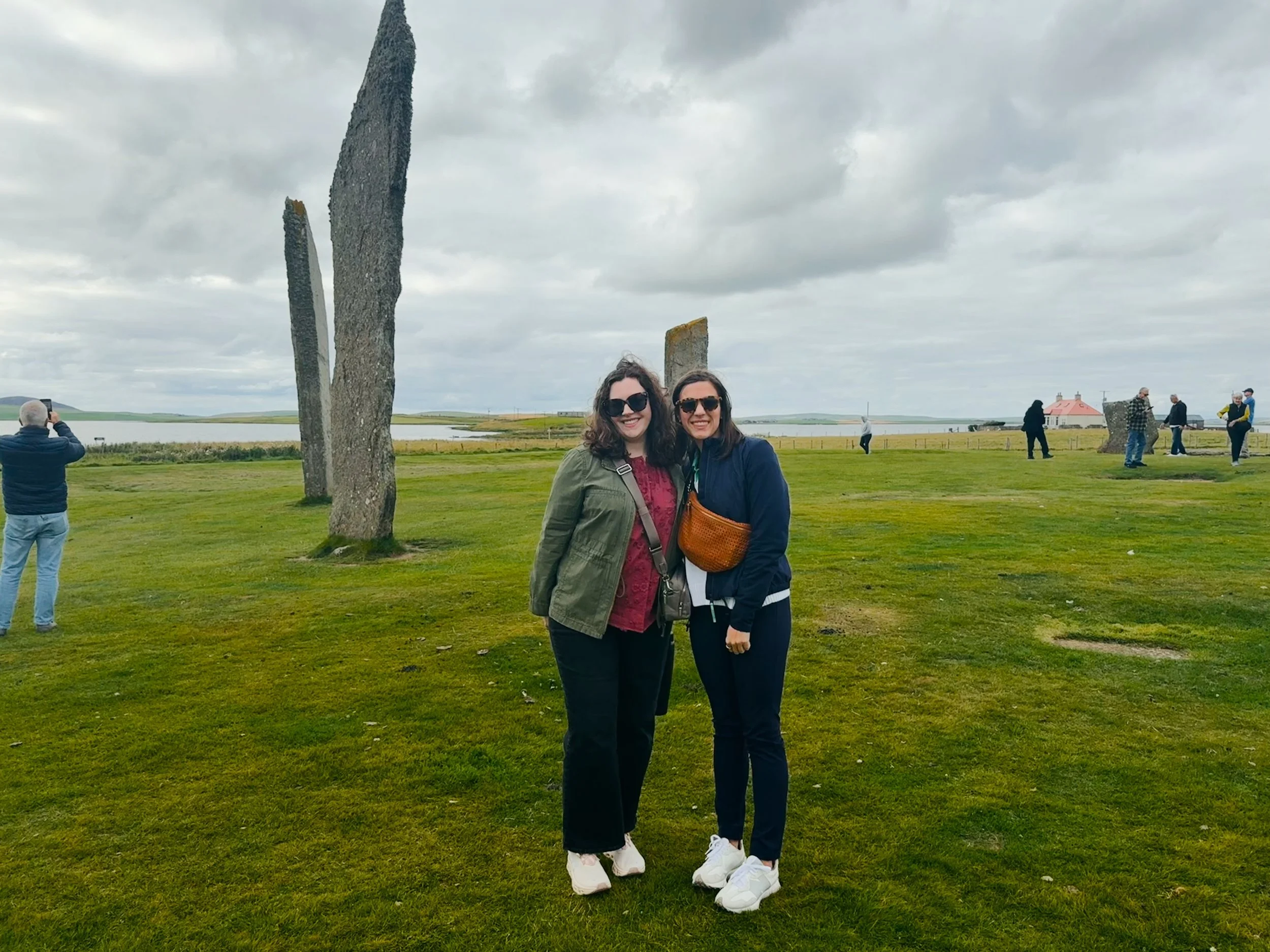 Two women standing together on a grassy field with large stone structures behind them, cloudy sky, and other people in the background, some taking photos, near a body of water and distant houses.