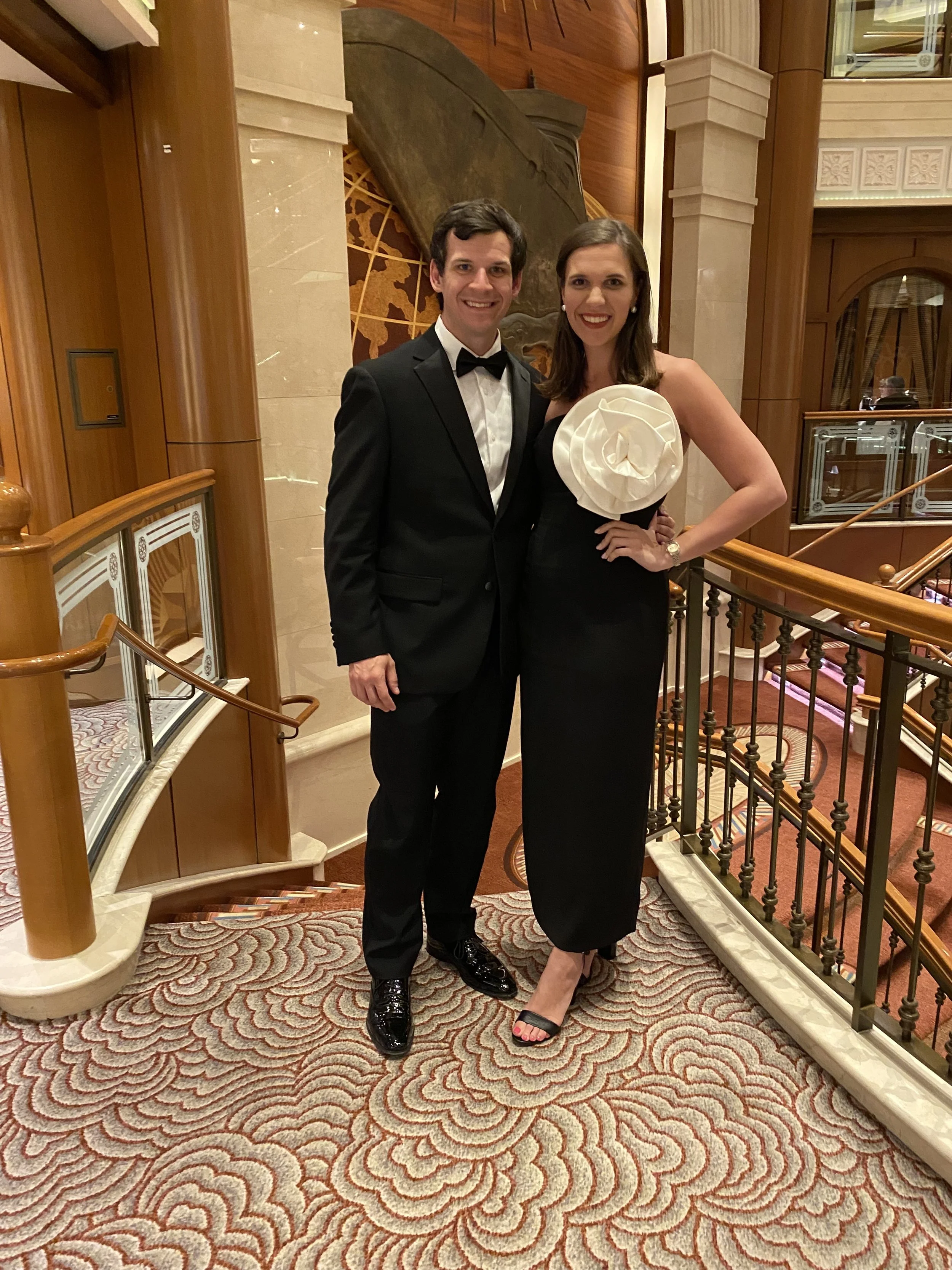 A man and woman dressed in tuxedo and evening gown, standing on a staircase, smiling at the camera in a fancy indoor setting.