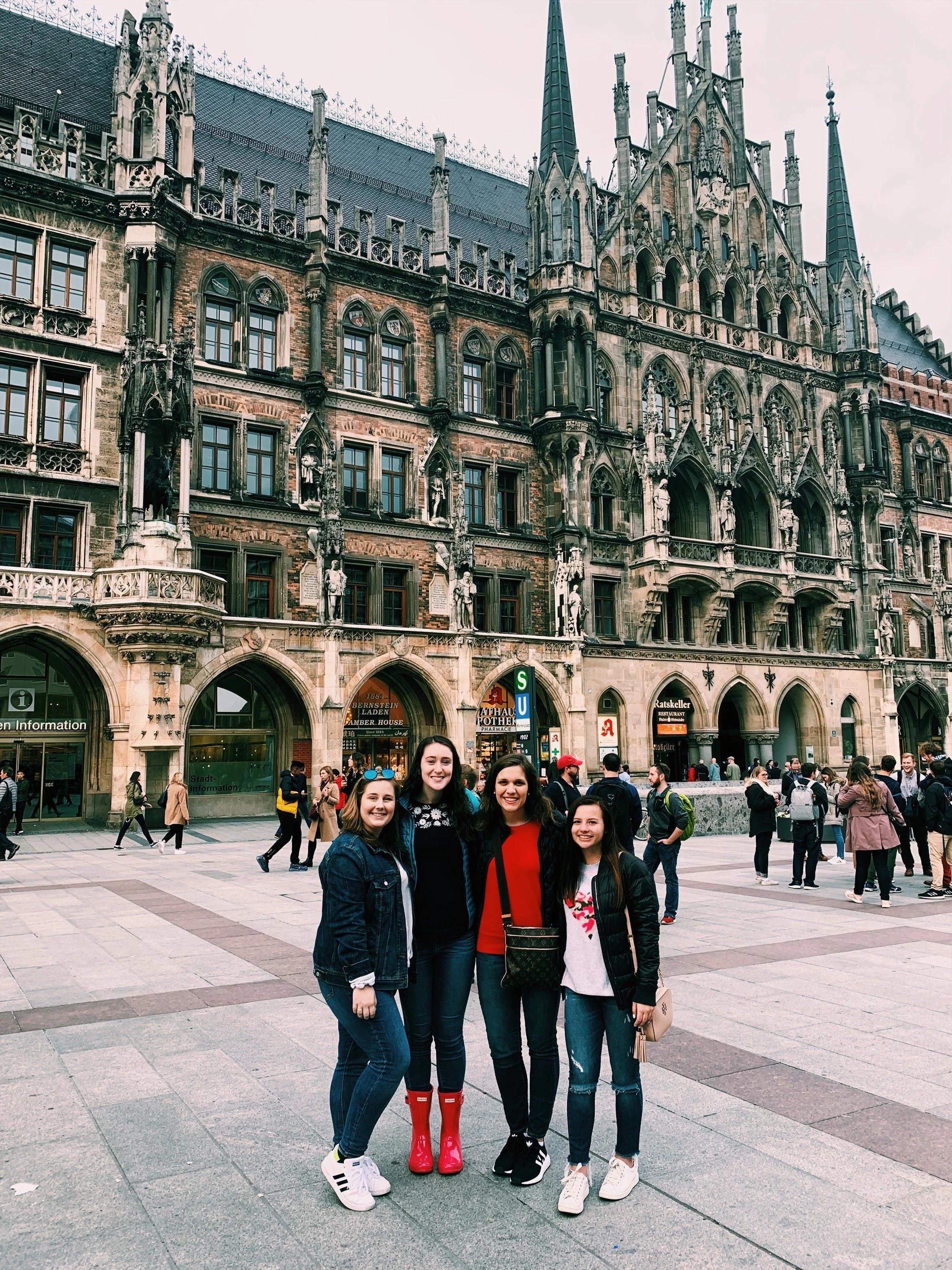 Four women smiling and posing together in a city square with a medieval-style building in the background, surrounded by other pedestrians.